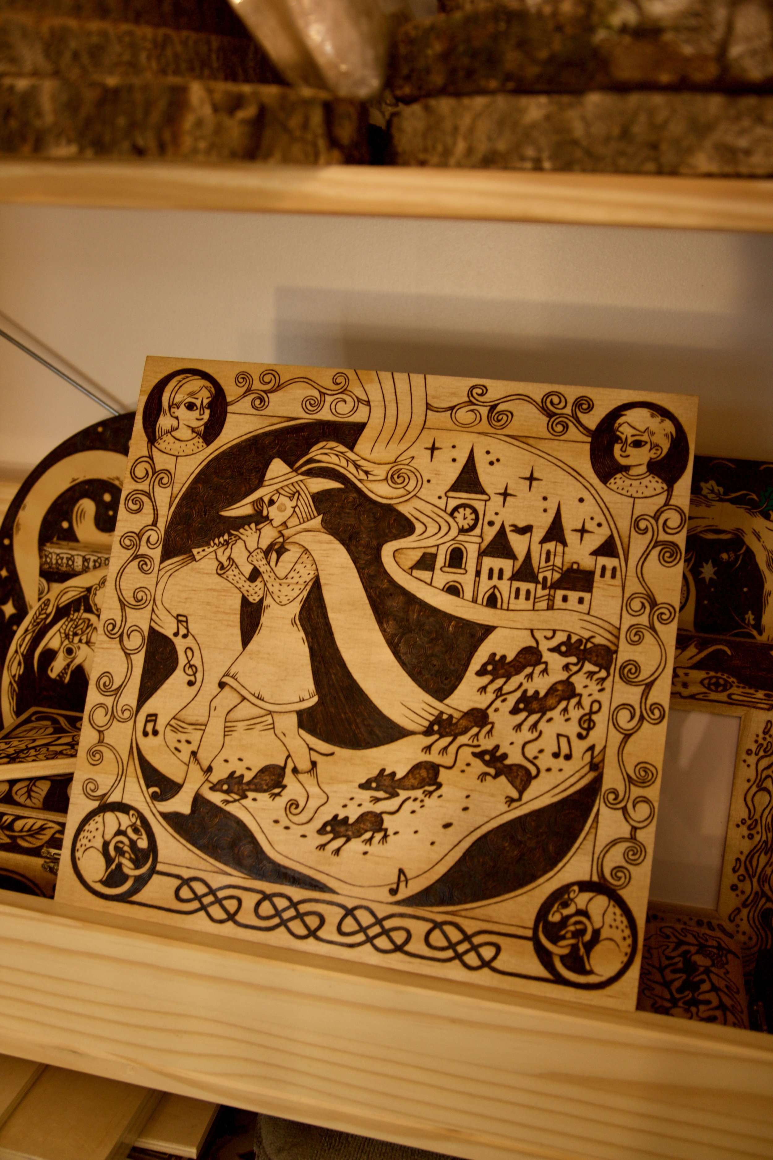 Vue entière d'une grande pyrogravure carrée sur bois représentant le joueur de flûte de Hamelin guidant les rats, entourée d'entrelacs médiévaux.