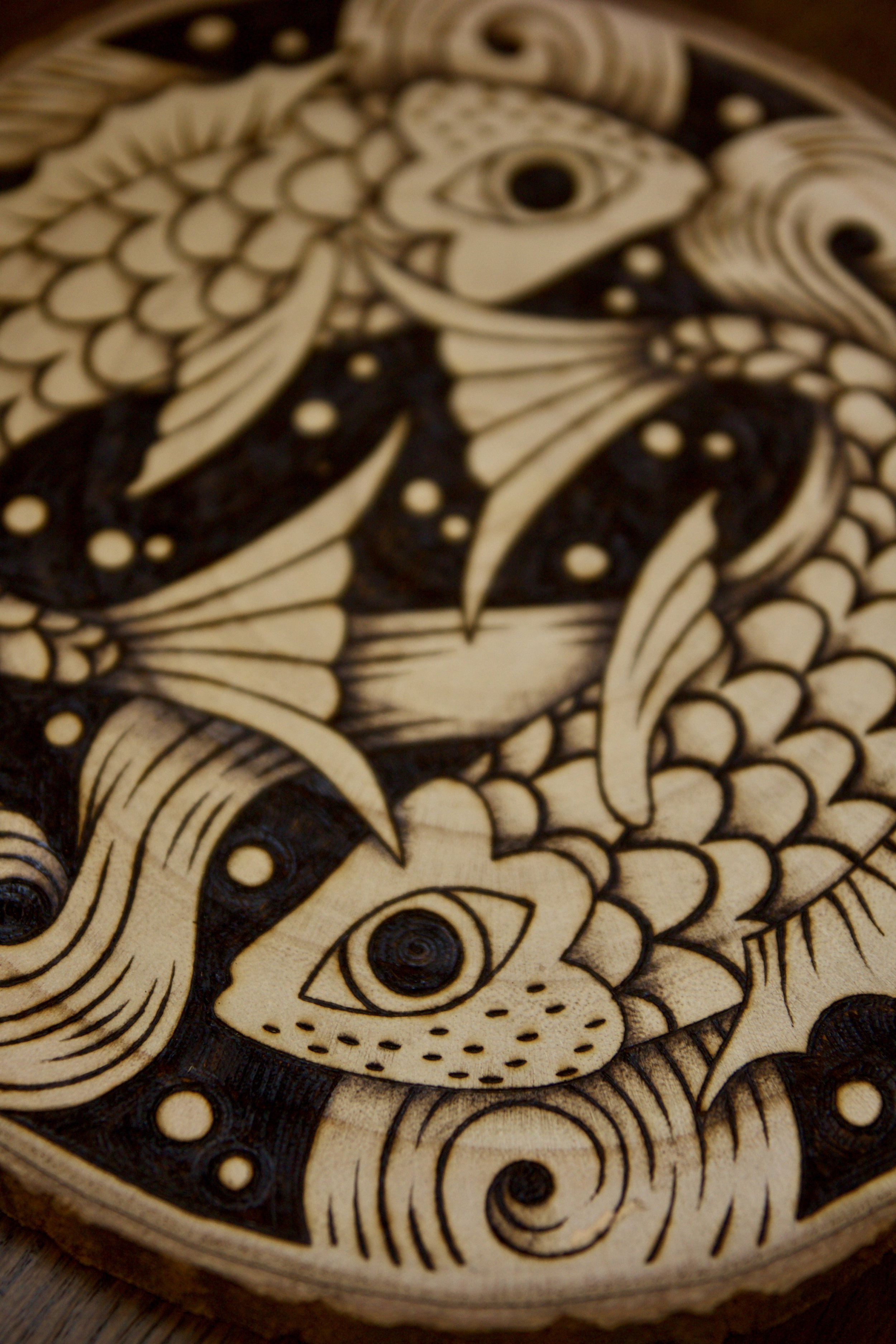 Détail d'une pyrogravure artisanale sur tranche de bois ronde représentant deux poissons entrelacés dans un style mystique et fluide évoquant le Yin Yang.