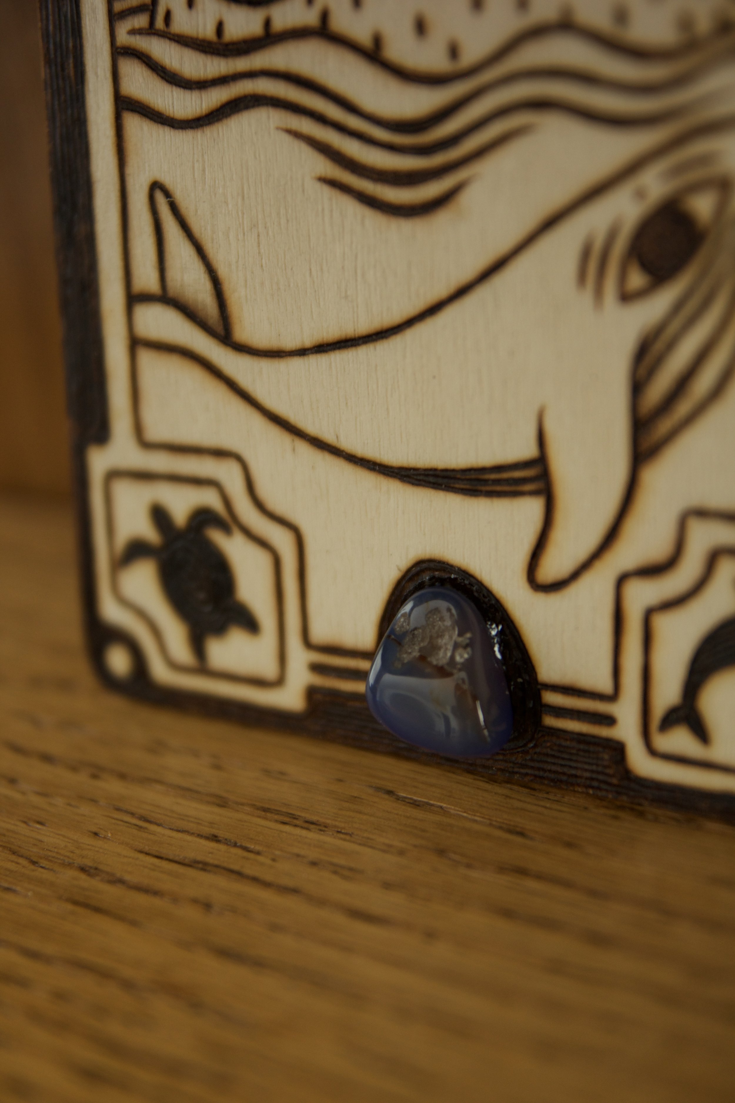 Zoom sur la pierre d'une petite plaque en bois gravée à la main de motifs marins et de vagues, sertie d'une pierre d'agate bleue naturelle aux reflets aquatiques.