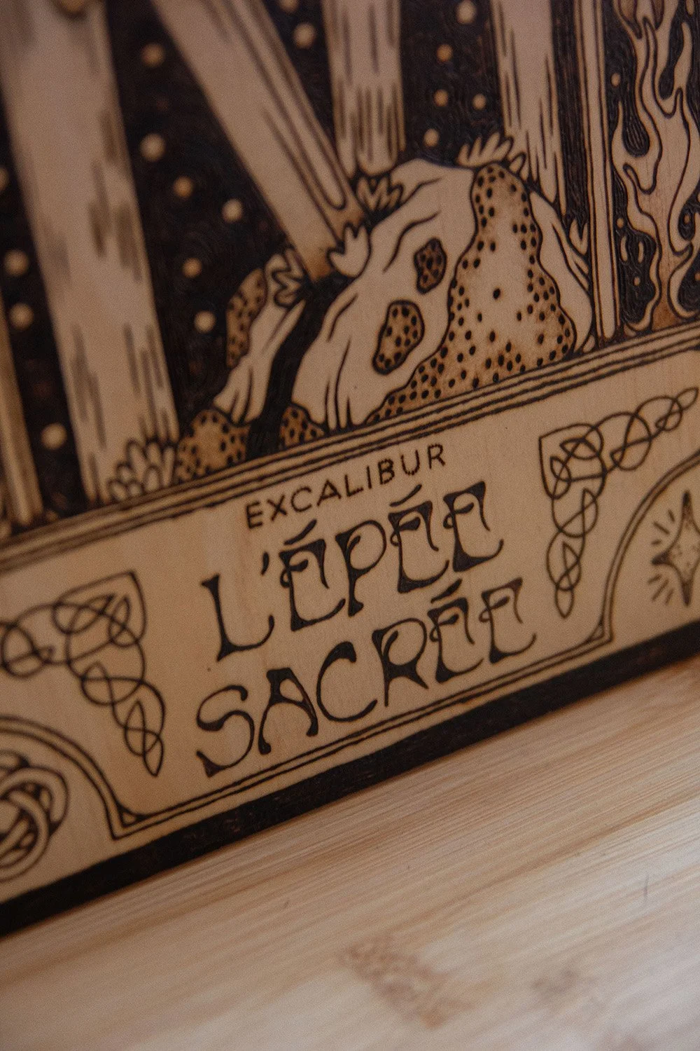 Zoom sur la typographie d'une pyrogravure artisanale faite main représentant l'épée sacrée Excalibur dans un style mystique et celtique, entourés de petites scènes et autres motifs folkloriques.