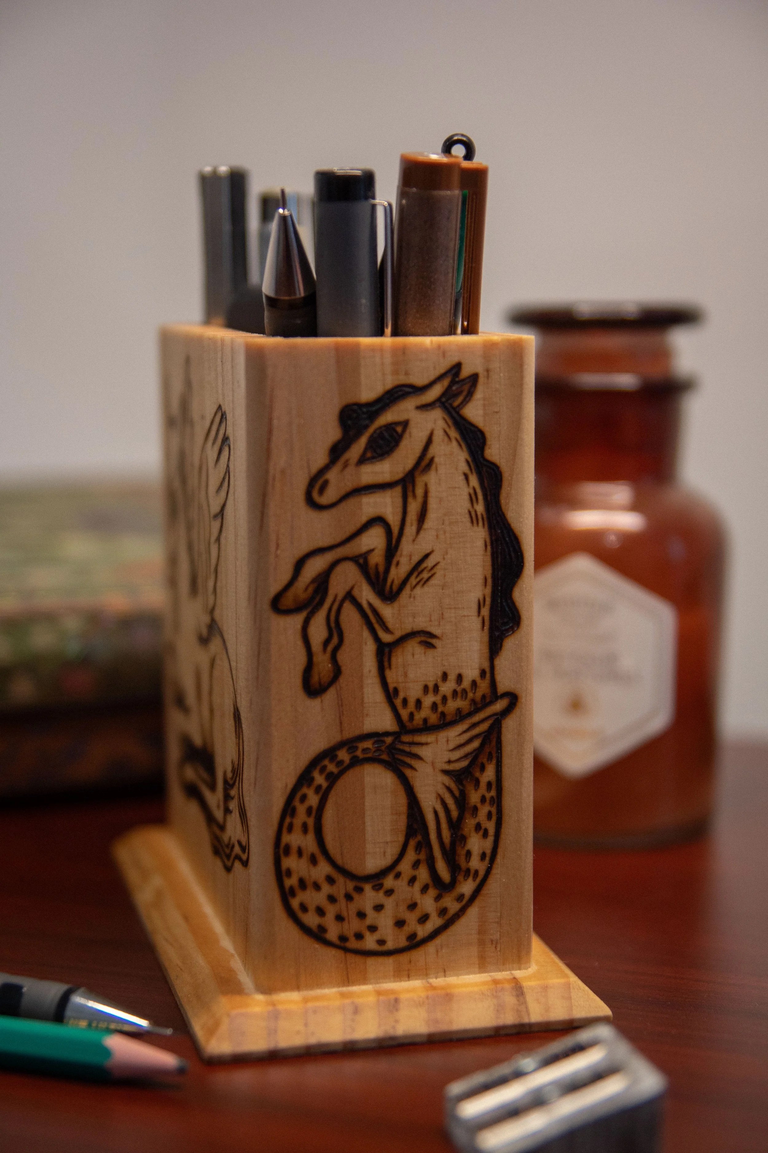 Vue de côté d'un pot à crayons carré en bois artisanal, montrant une face pyrogravée avec soin représentant un hippocampe magique.