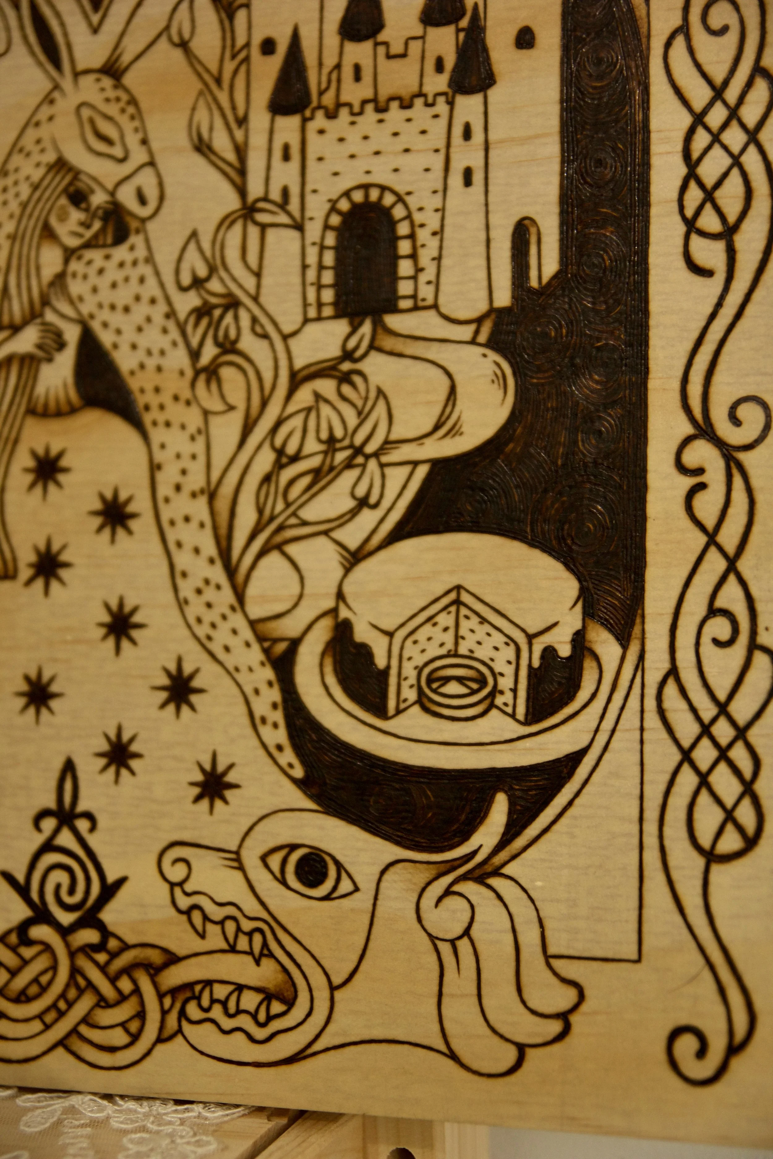 Zoom sur l'anneau tombé dans un gateau d'un tableau carré en bois pyrogravé à la main représentant une illustration du conte Peau d’Âne inspirée du style des enluminures médiévales.