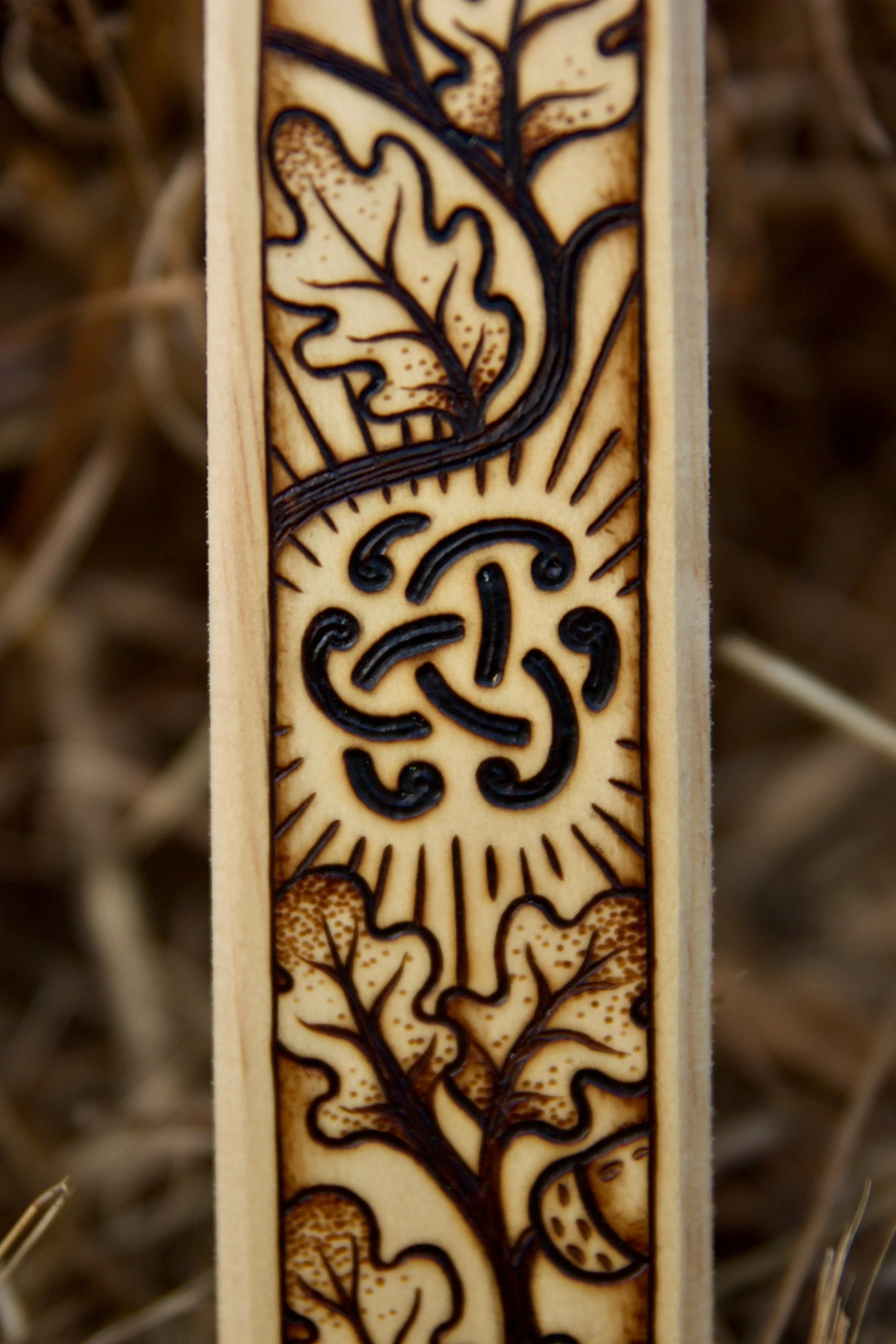Détail motif magique sur une épée en bois artisanale plantée dans des herbes folles sauvages, finement pyrogravée avec des nœuds celtiques sur la garde et des motifs de glands et de chêne sur la lame.