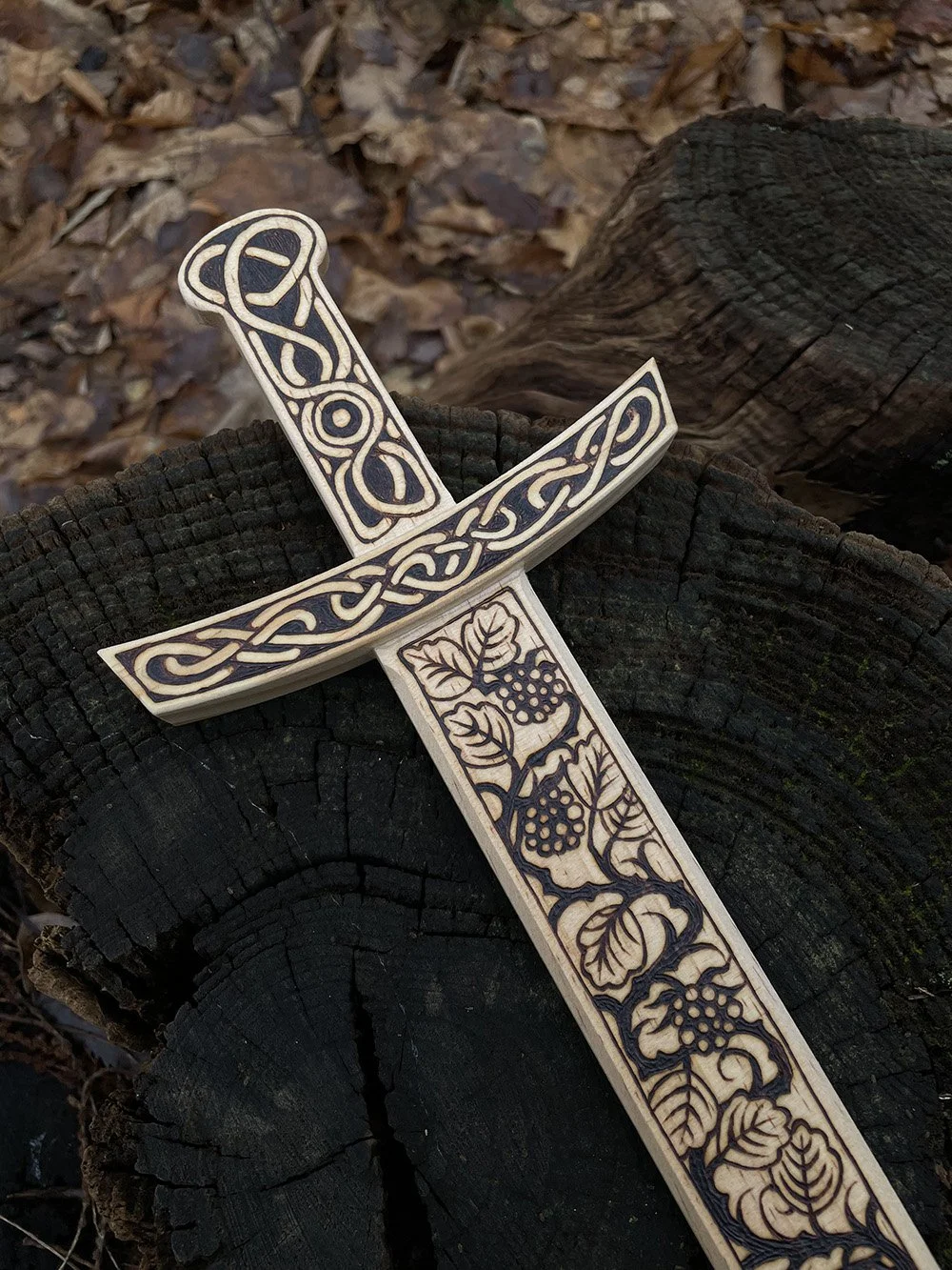 Épée décorative en bois clair posée sur un tronc d'arbre entièrement pyrogravée à la main avec des motifs d'entrelacs celtiques, nœuds mystiques et inspirations médiévales.
