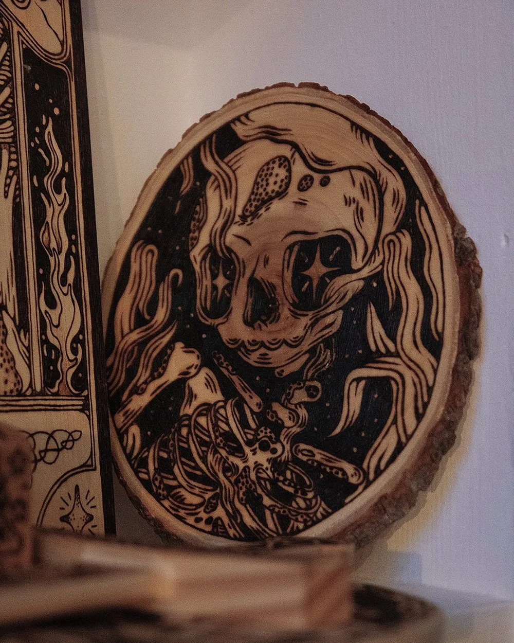 Pyrogravure artisanale sur bois brut ovale représentant un squelette Memento Mori au style d'un artefact ancien présentée sur une étagère.