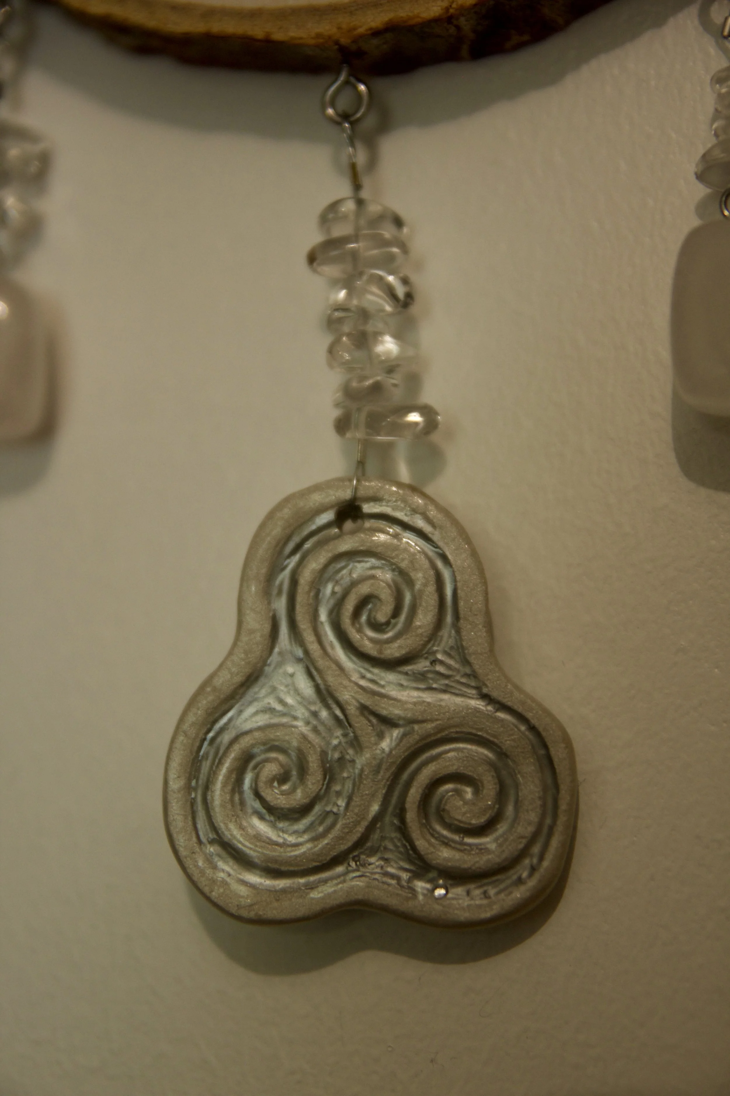 Élément en céramique grise faite main représentant une triskèle relié à une amulette en bois par des perles en perles précieuses.
