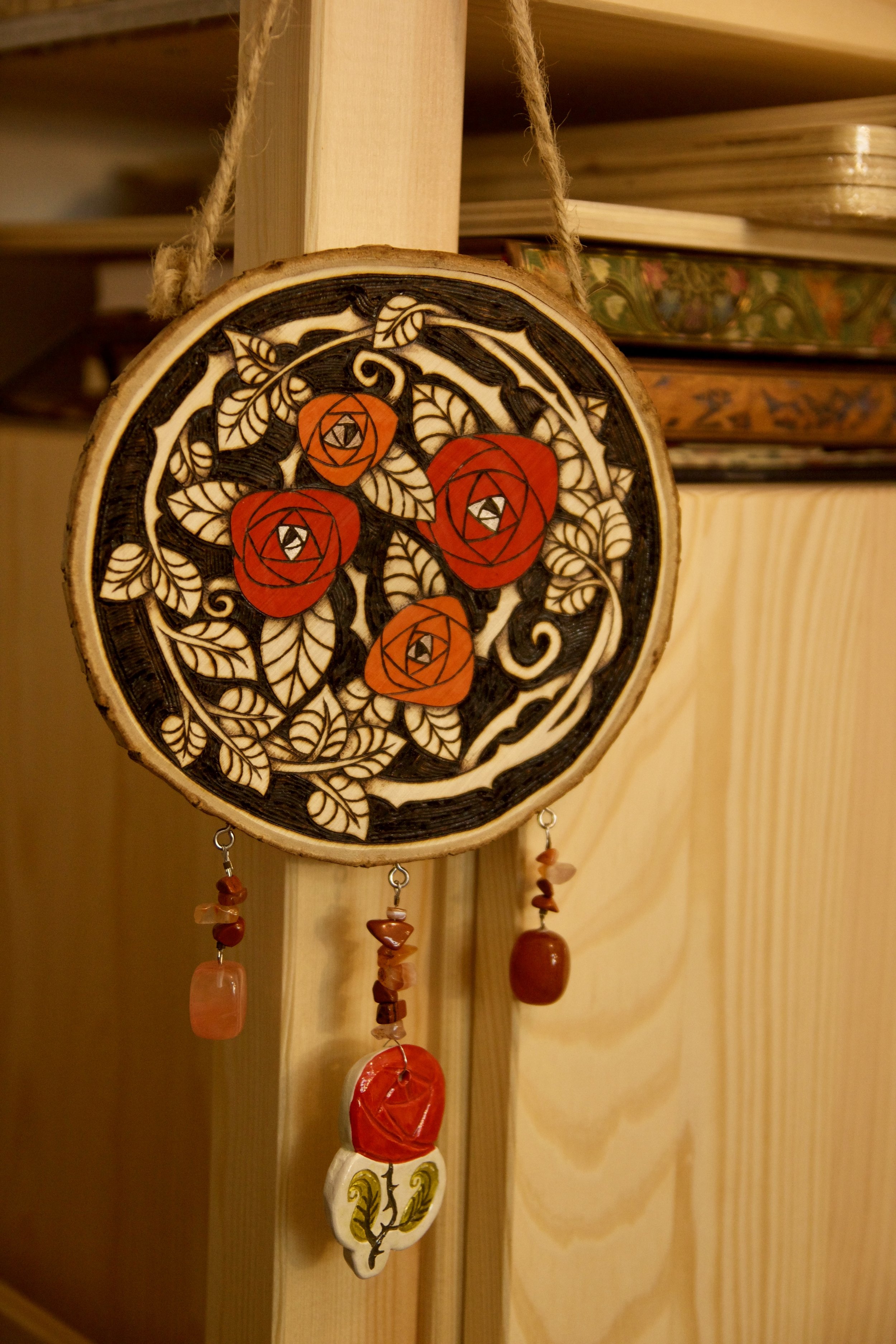 Zoom sur une amulette murale ronde en bois pyrogravé et peint avec des roses rouges, ornée de pendants en pierre et d'une rose en céramique.