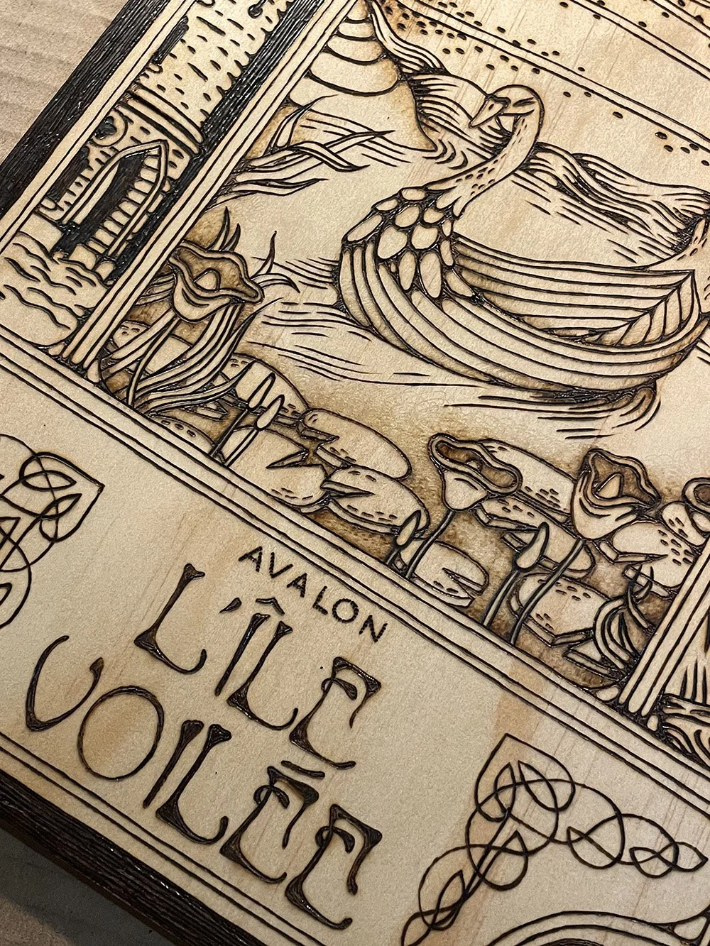 Détail d'une pyrogravure artisanale sur planche de bois évoquant une illustration ancienne de l'île mythique d'Avalon dans la brume.