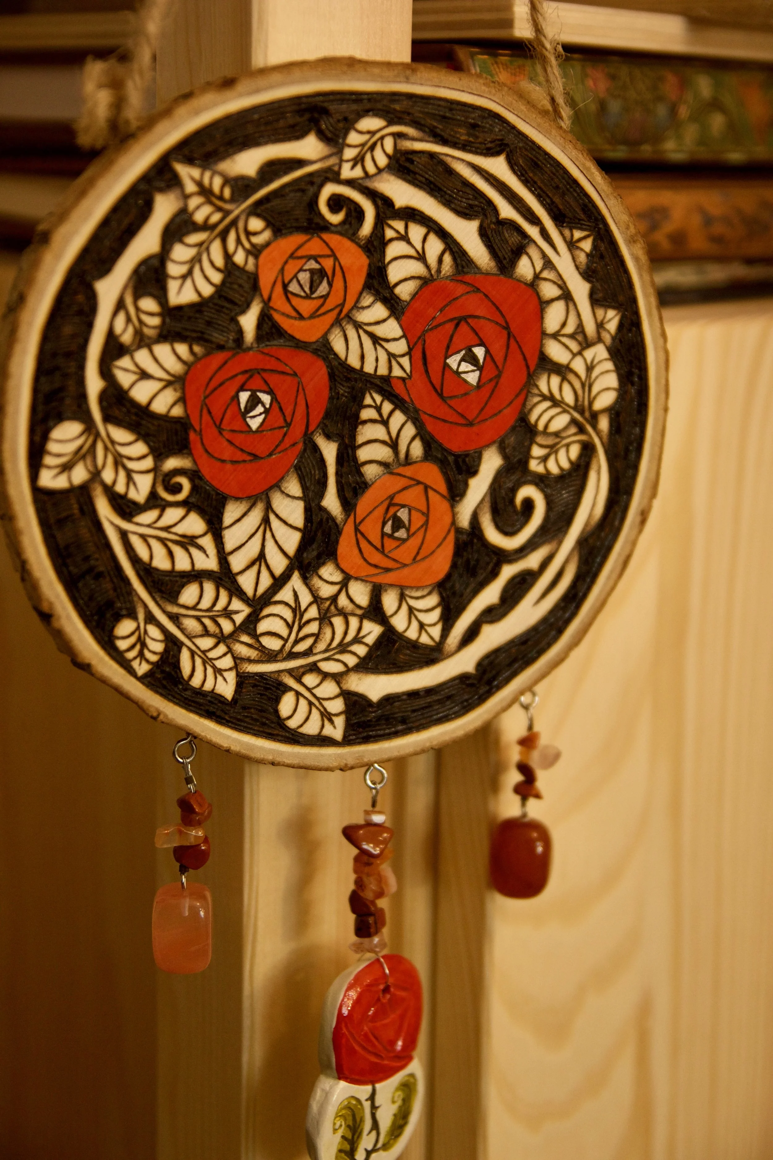 Détail sur une amulette murale ronde en bois pyrogravé et peint avec des roses rouges, ornée de pendants en pierre et d'une rose en céramique.