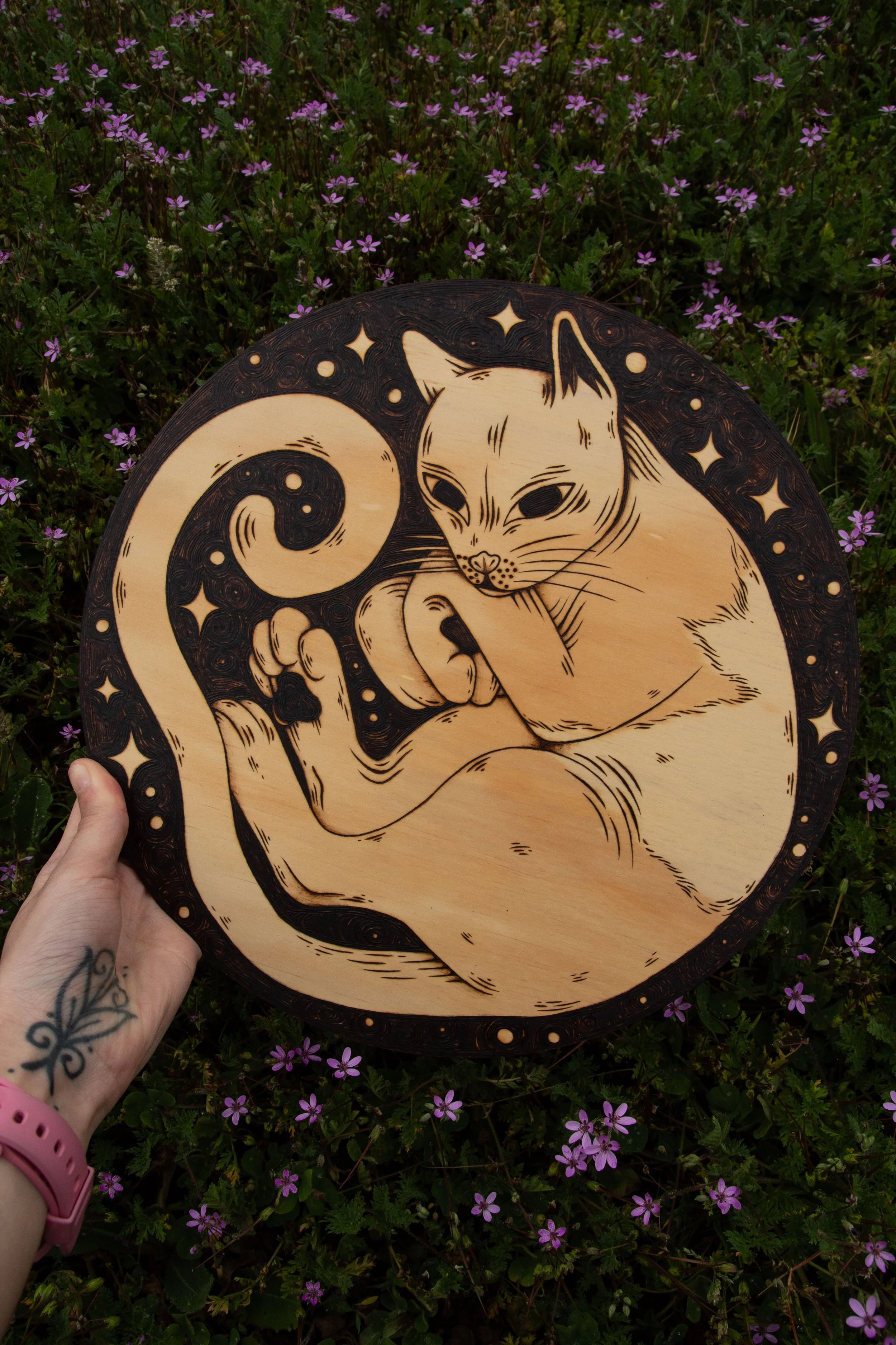 Pyrogravure artisanale sur un disque en bois naturel représentant un chat mystique gravé avec de fins détails de pelage et d'ombrage dans un parterre de fleurs violettes.