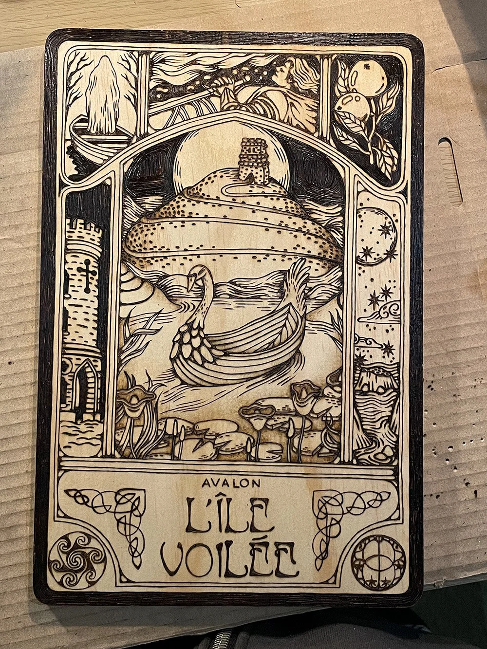 Pyrogravure artisanale sur planche de bois évoquant une illustration ancienne de l'île mythique d'Avalon dans la brume dans un atelier d'artiste.