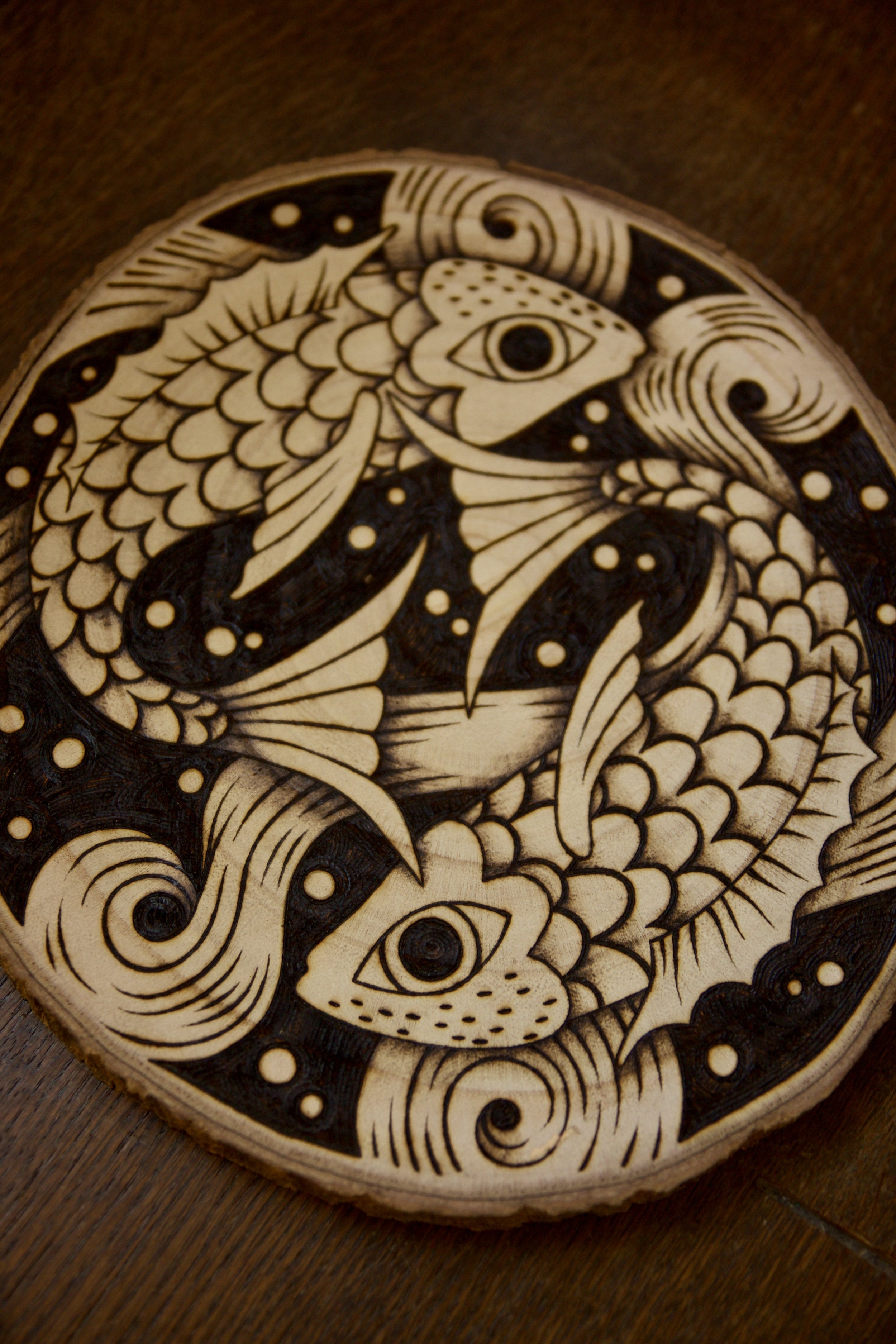 Zoom sur une pyrogravure artisanale sur tranche de bois ronde représentant deux poissons entrelacés dans un style mystique et fluide évoquant le Yin Yang.