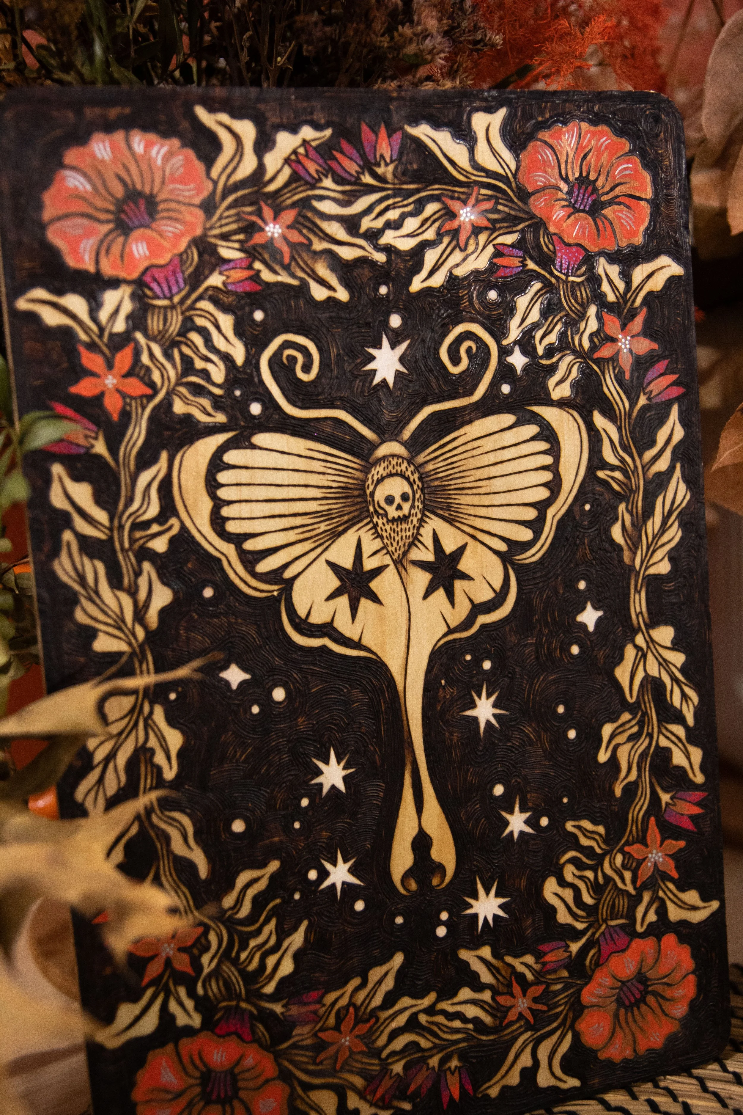 Zoom sur une plaque en bois artisanale pyrogravée d'un papillon de nuit mystique entouré de fleurs délicates sublimées par des touches de peinture colorée.