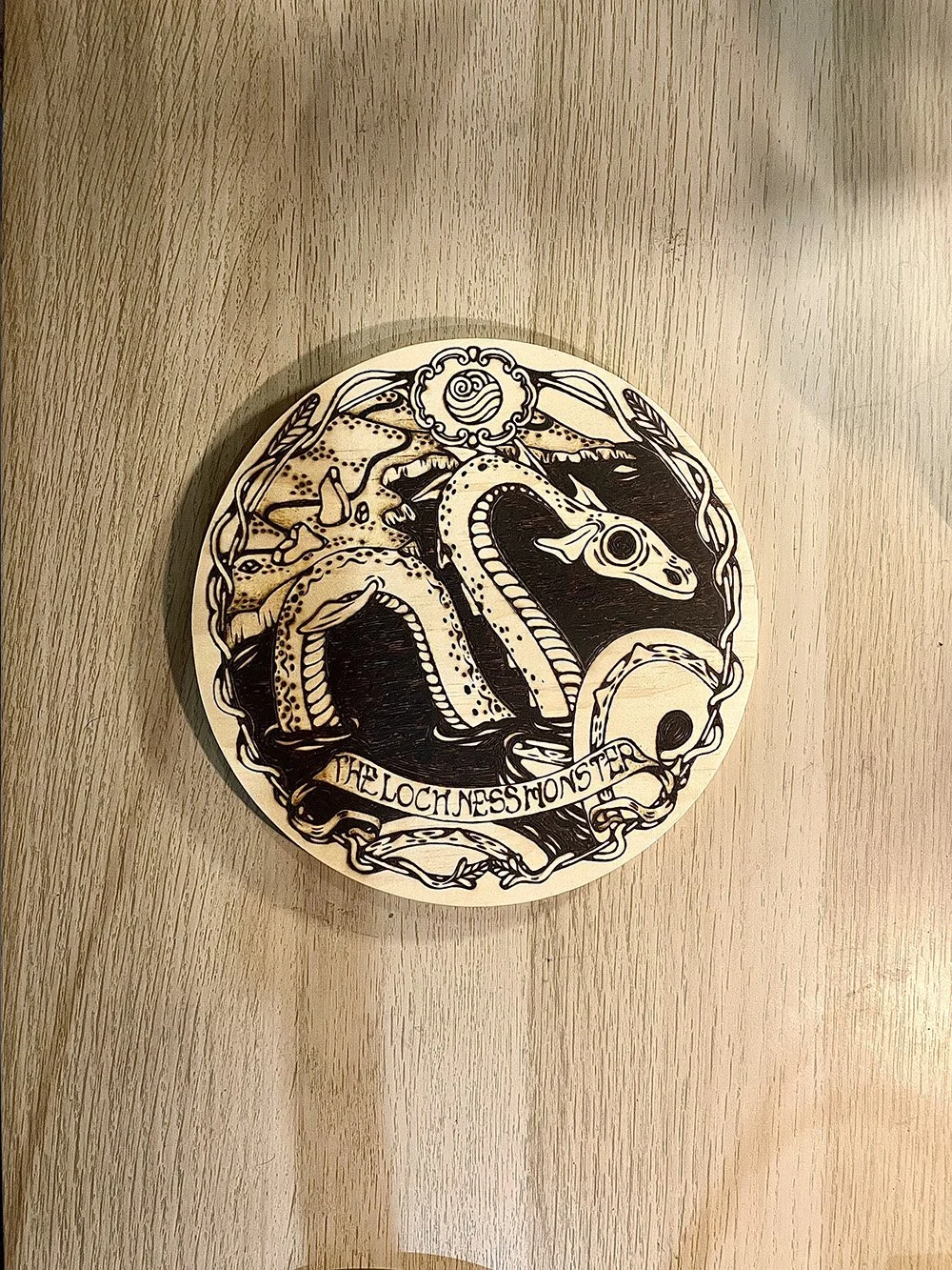 Pyrogravure artisanale ronde sur bois représentant la silhouette mystique du monstre du Loch Ness sous les eaux sombres d'Écosse sur un bureau d'atelier d'artiste.