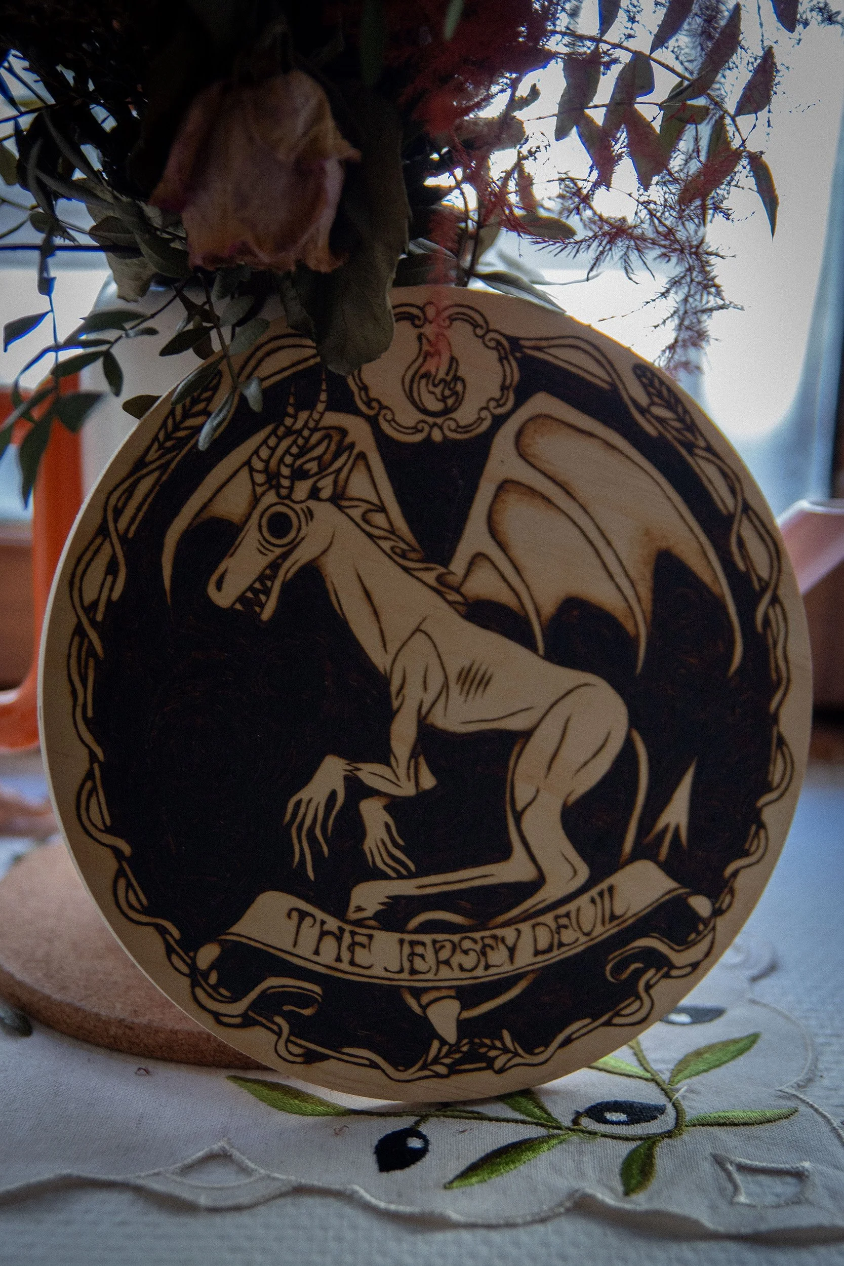 Pyrogravure artisanale ronde de 20x20 cm sur bois représentant la silhouette du Diable de Jersey, créature ailée mythique du folklore américain dans des teintes orangées.