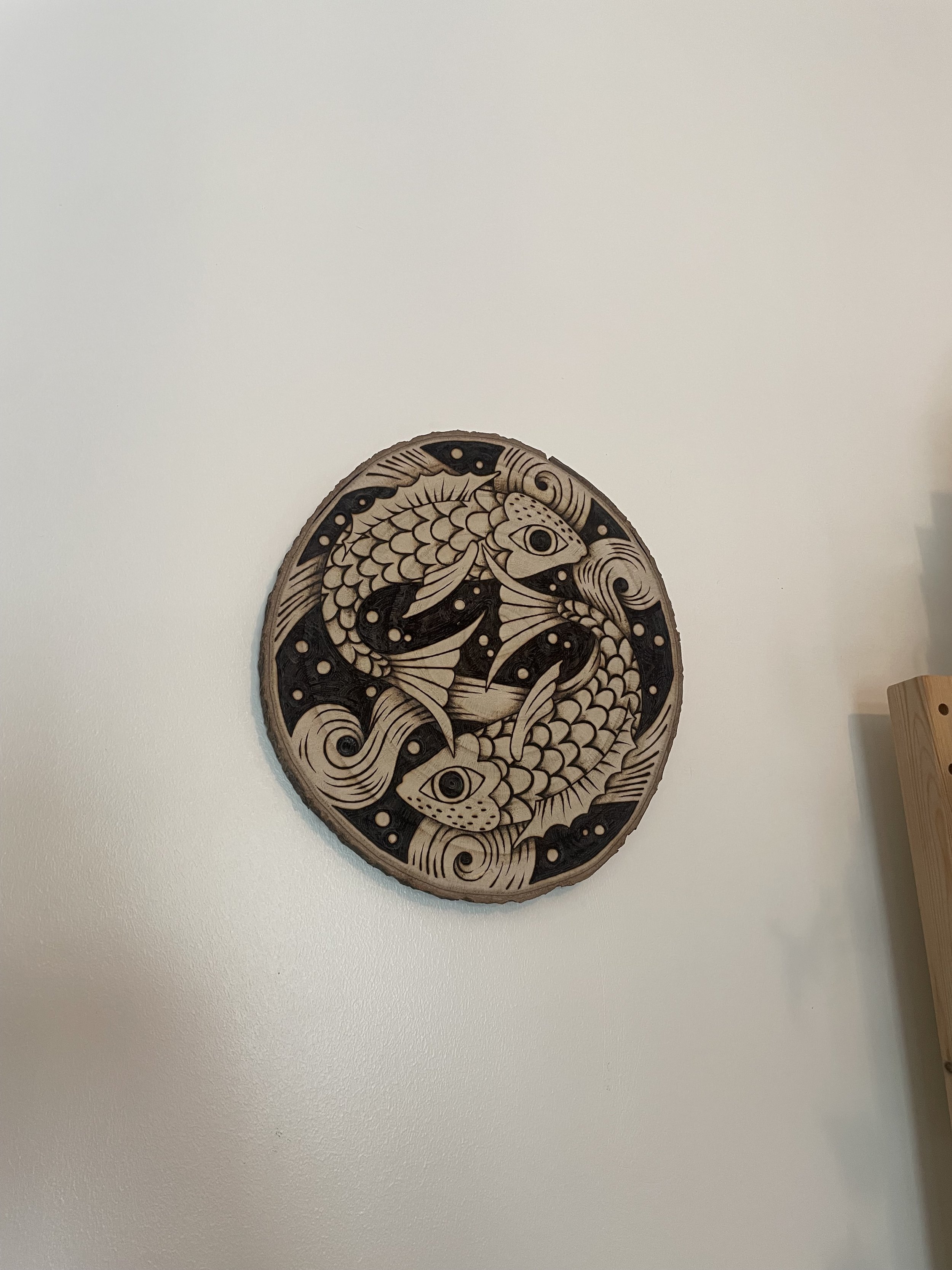 Pyrogravure artisanale représentant deux poissons entrelacés accroché sur un mur blanc.