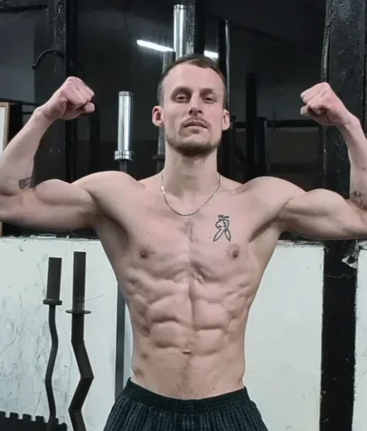 A shirtless man flexing his biceps in a gym with a serious expression.