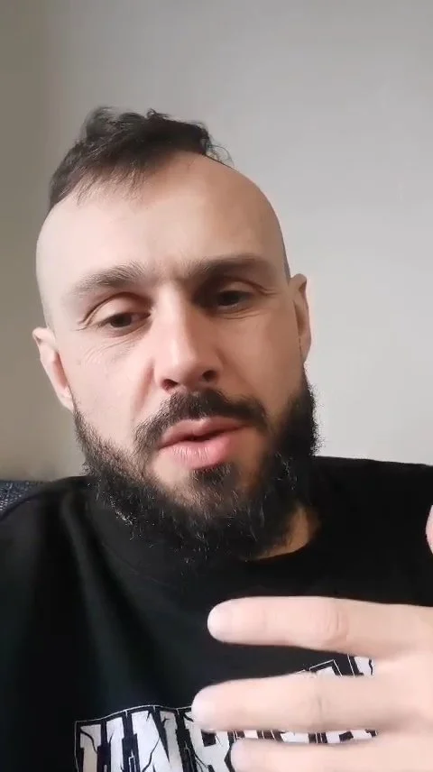 A man with a beard, short hair, and a black shirt pointing with his finger, indoors against a plain light-colored wall.