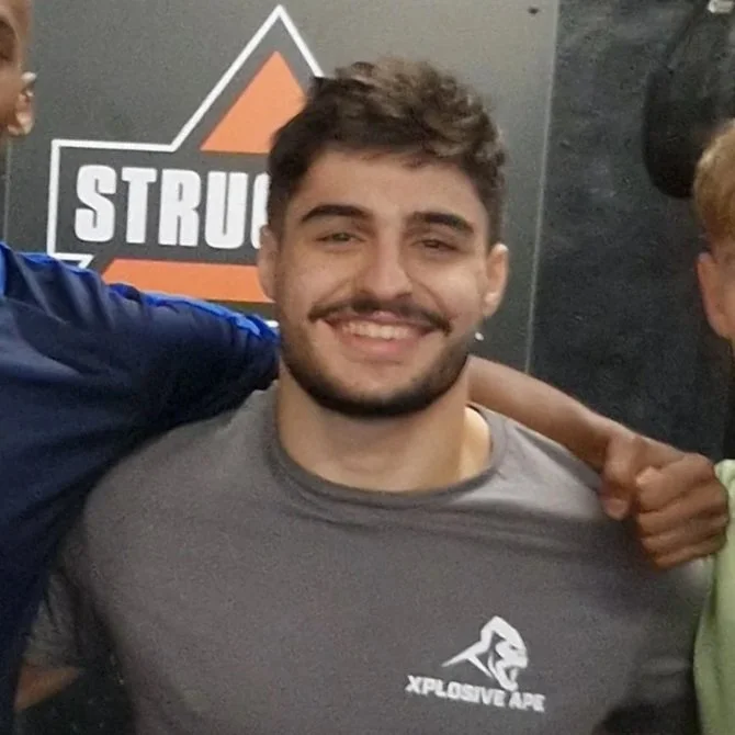 Young man with short brown hair and a beard smiling, wearing a gray T-shirt with 'Xplosive Ape' logo, standing in front of a black and gray wall with a partially visible logo that says 'STRU'.
