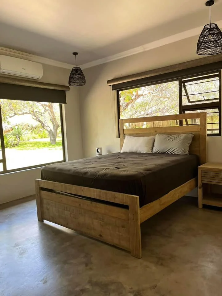 A bedroom with a wooden bed, dark bedding, and two pillows. Large windows with blinds show a view of greenery outside. Two black hanging light fixtures are on the ceiling, and a small nightstand is beside the bed.