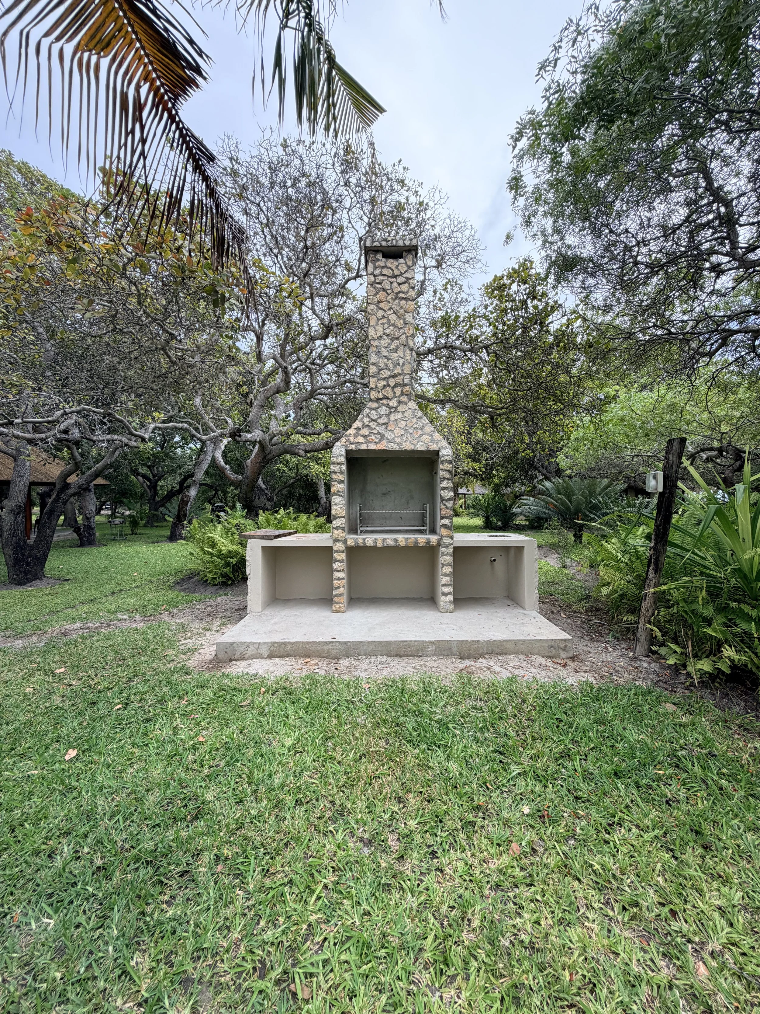 A stone outdoor fireplace with a chimney in a lush green backyard, surrounded by trees and plants.