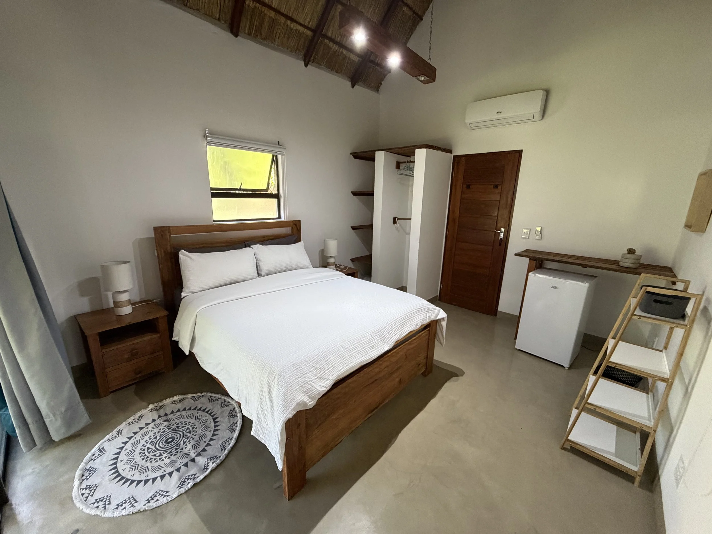 A bedroom with a wooden bed frame and white bedding, a nightstand with a lamp, a round patterned rug, a small white refrigerator, a folding towel rack, a wooden door, a window, and a wall-mounted air conditioner.
