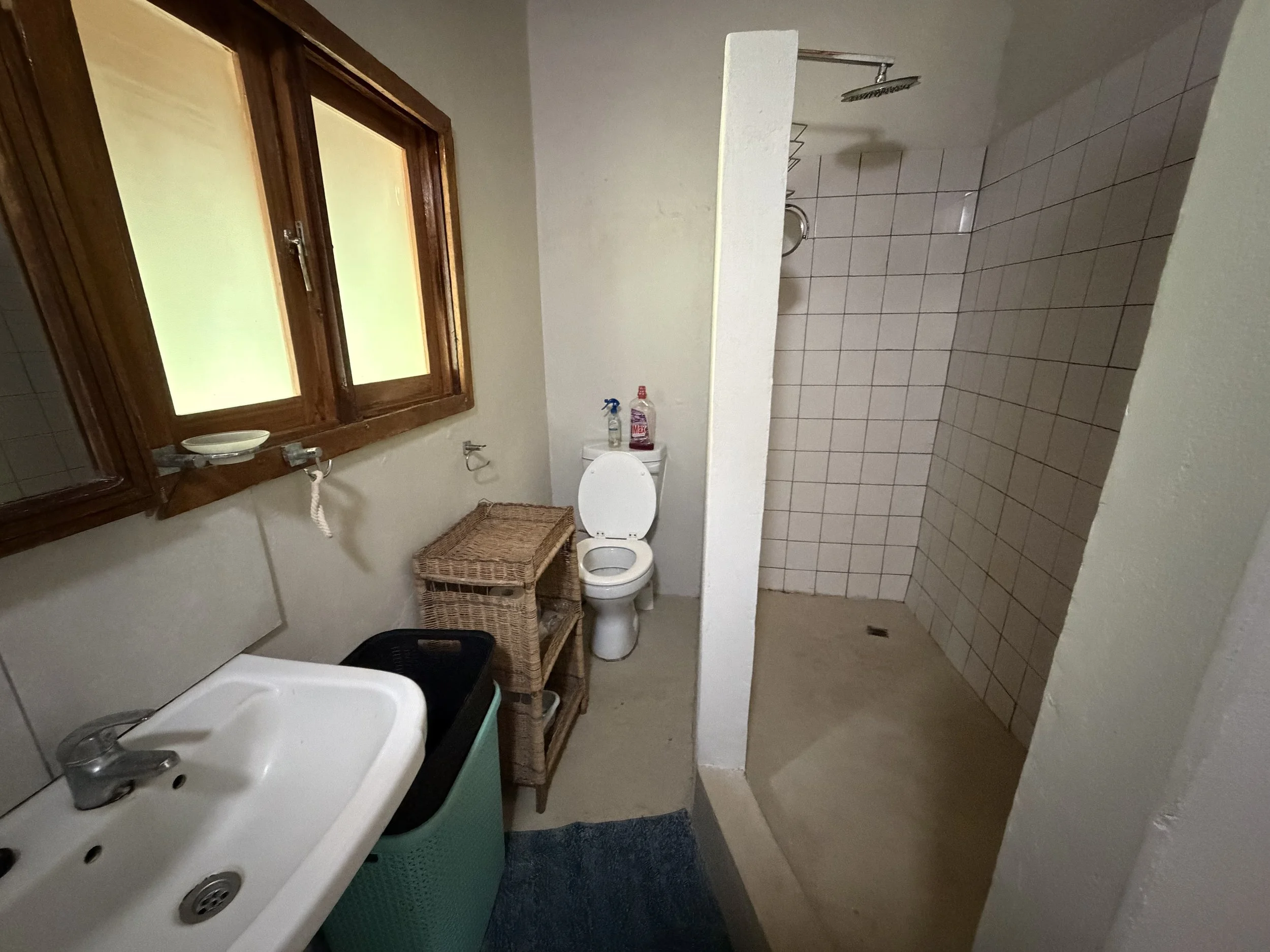 A bathroom with a small sink in the foreground, a shower area with tiled walls to the right, a toilet with cleaning supplies on top of the tank, a wicker basket, and a wooden window with frosted glass on the left side of the wall.