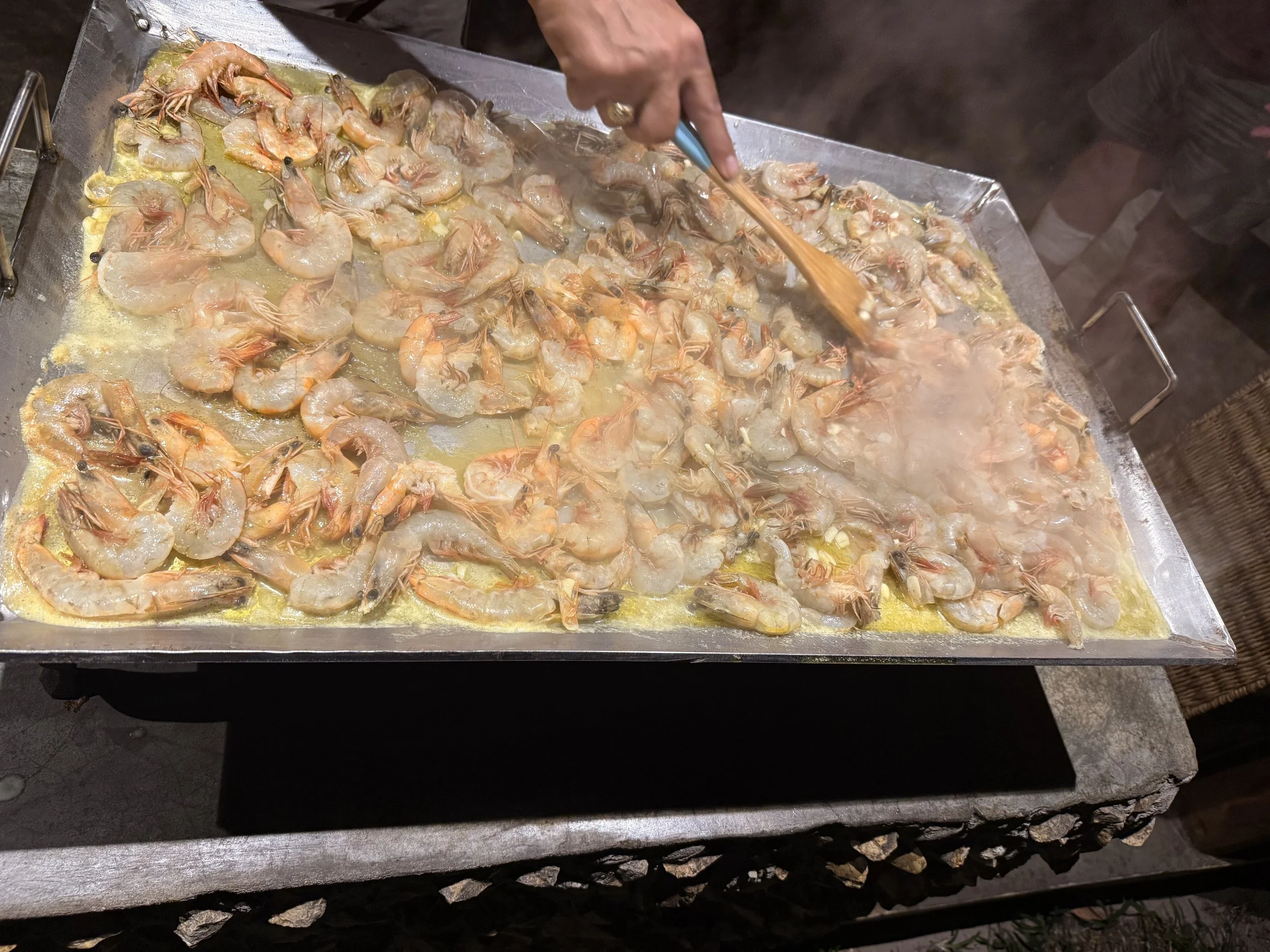 A person cooking shrimp on a flat grill, stirring with a wooden spatula, with steam rising from the hot shrimp.