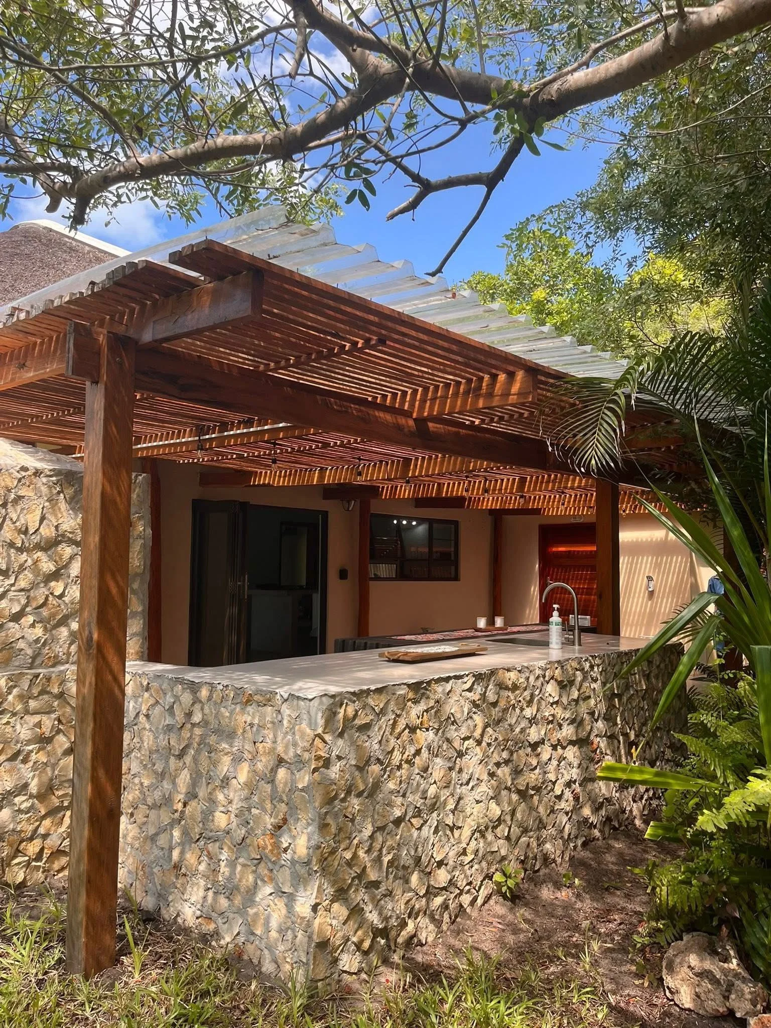 An outdoor kitchen with a stone countertop and a wooden pergola, surrounded by green plants and trees with a blue sky overhead.