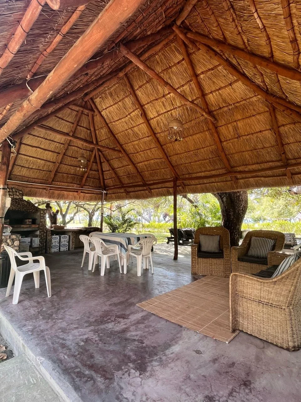 Open-air thatched roof pavilion with wooden beams, plastic and wicker chairs, a table with a tablecloth, and surrounded by trees and greenery.