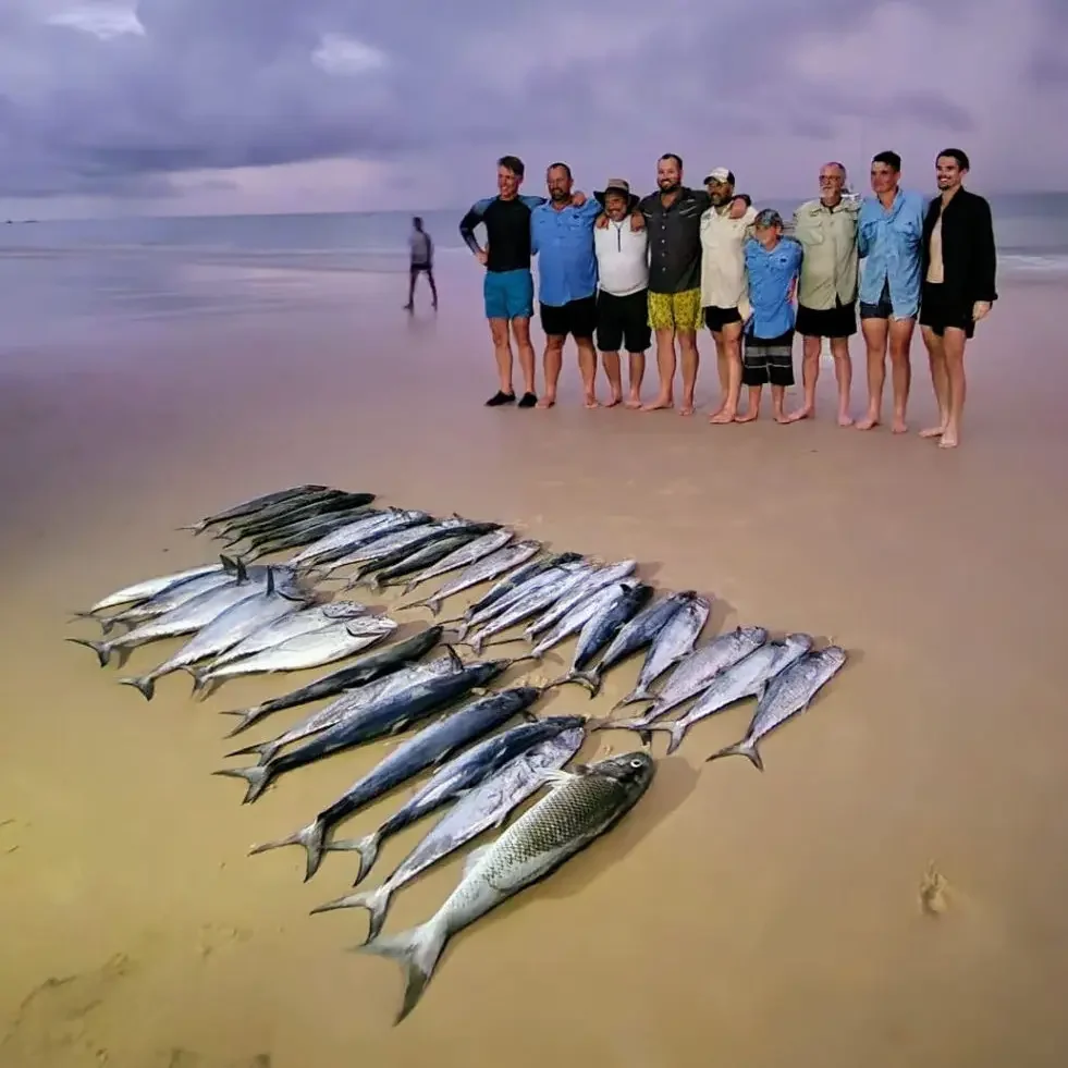 A group of people standing on the beach near the ocean, with a large catch of fish laid out in front of them. The sky is cloudy, and some of the people are wearing beachwear and hats.