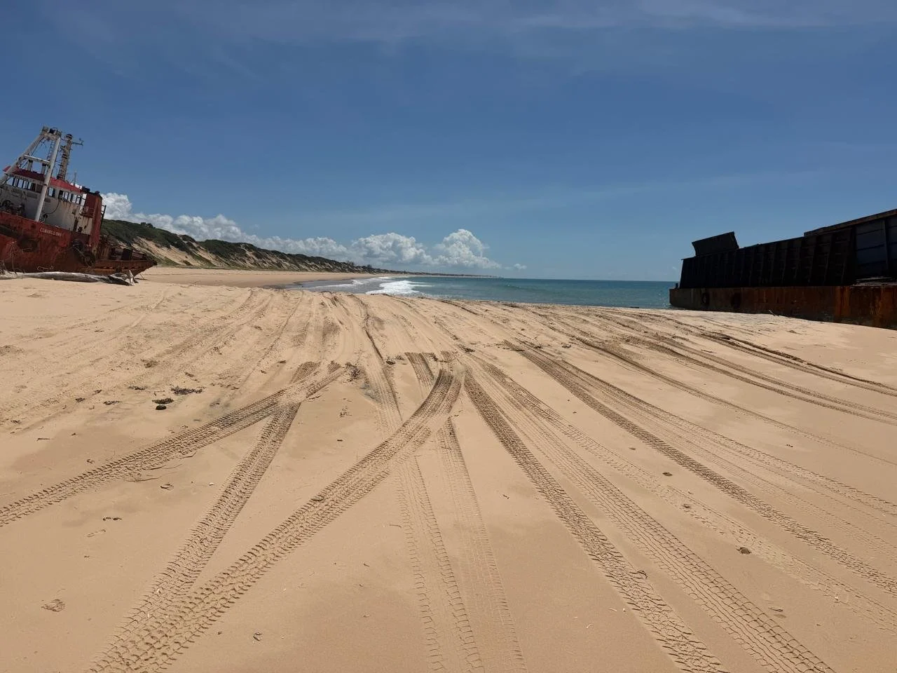 The image shows a deserted sandy beach with clear blue sky, scattered clouds, and tire tracks in the sand. Old, rusted ships are stranded on the shore on both sides of the beach, and distant dunes are visible in the background.