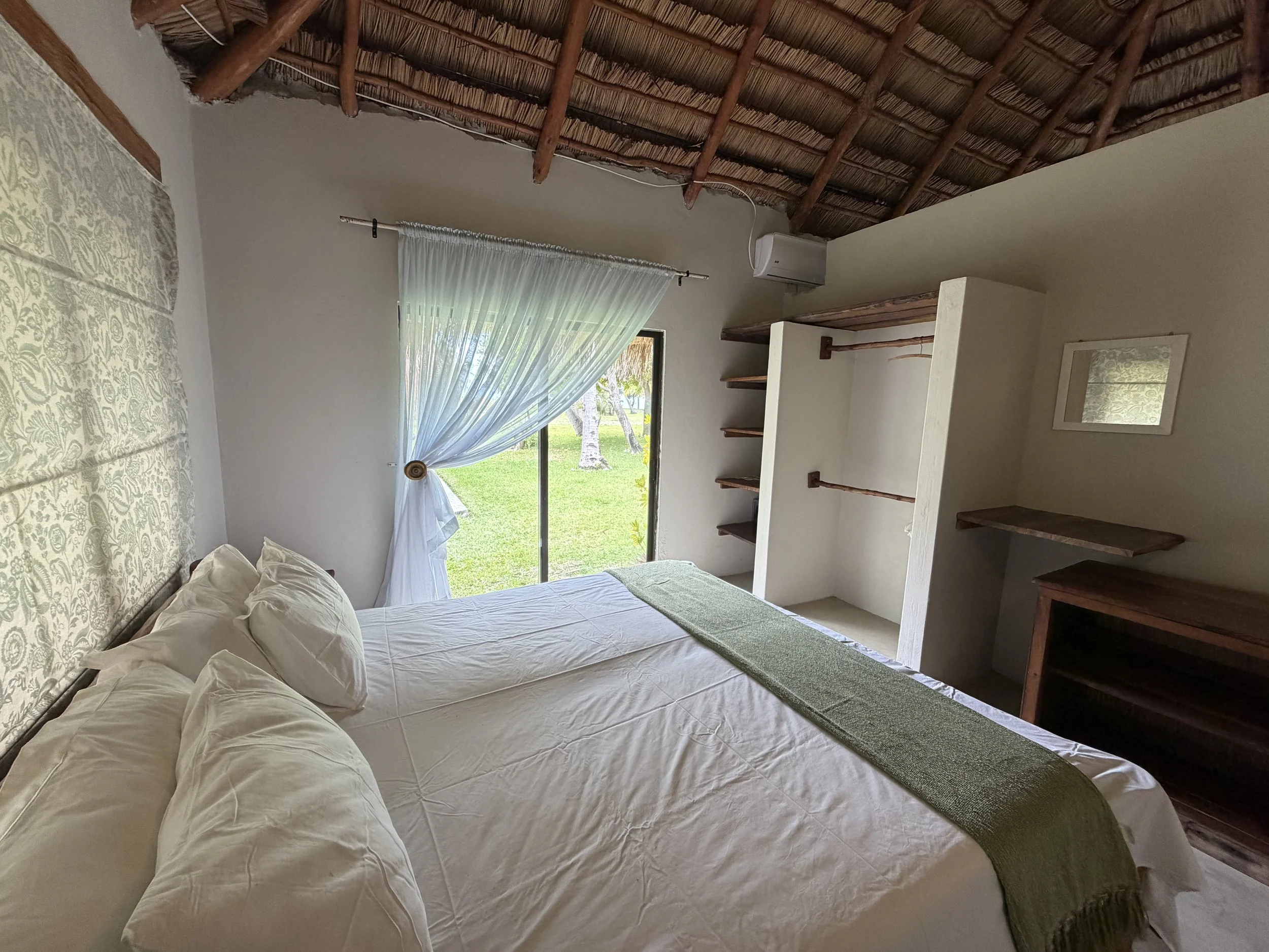A cozy bedroom with a large bed covered in white linens and pillows, a window with sheer curtains opening to a green outdoor scene, wooden shelves, and a thatched roof ceiling.