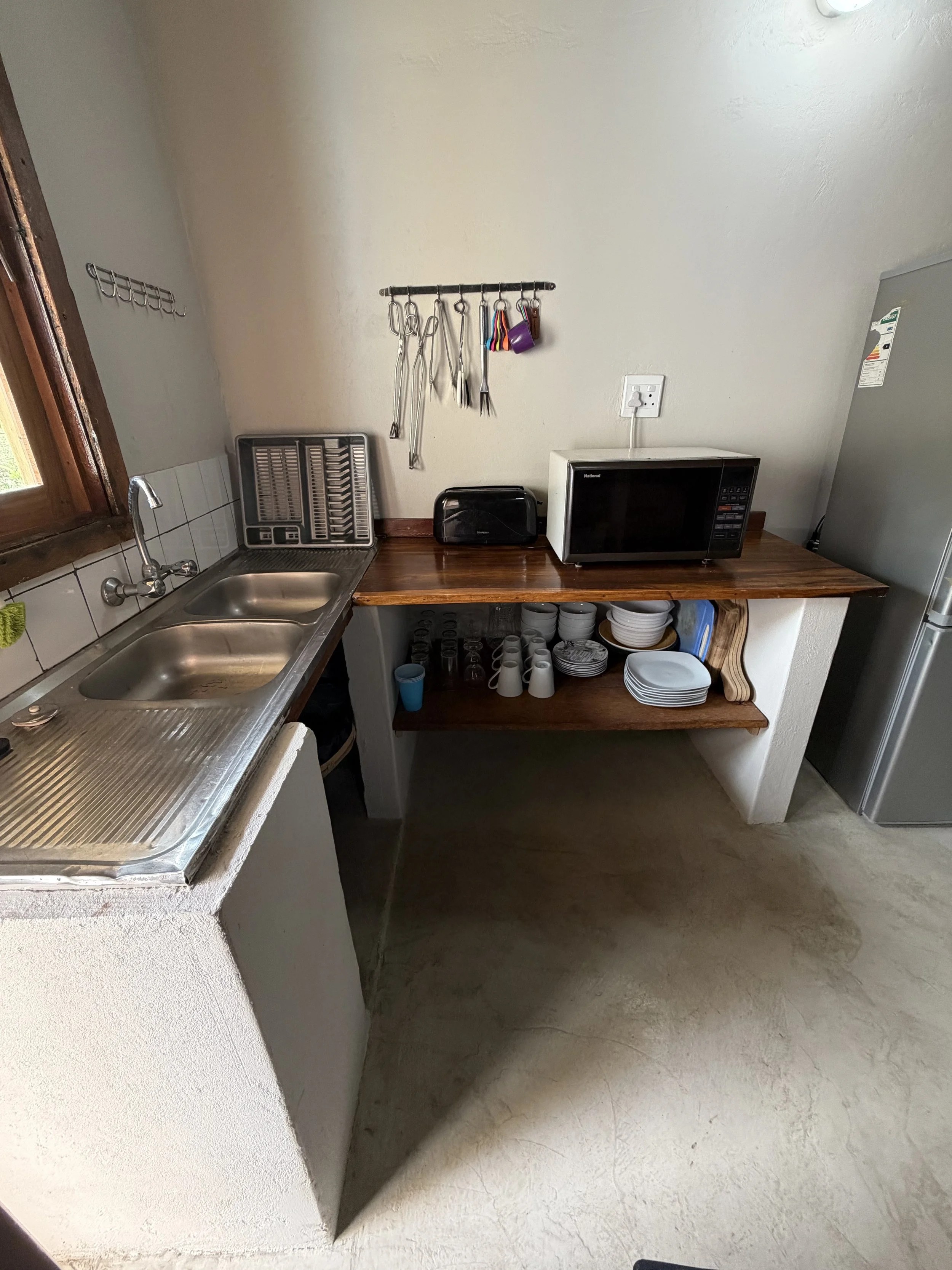 A small kitchen counter with a double sink, a dish drying rack, a microwave oven, a toaster, and a cutting board. Above the counter, a wall-mounted rack holds kitchen utensils, and a window with a wooden frame.