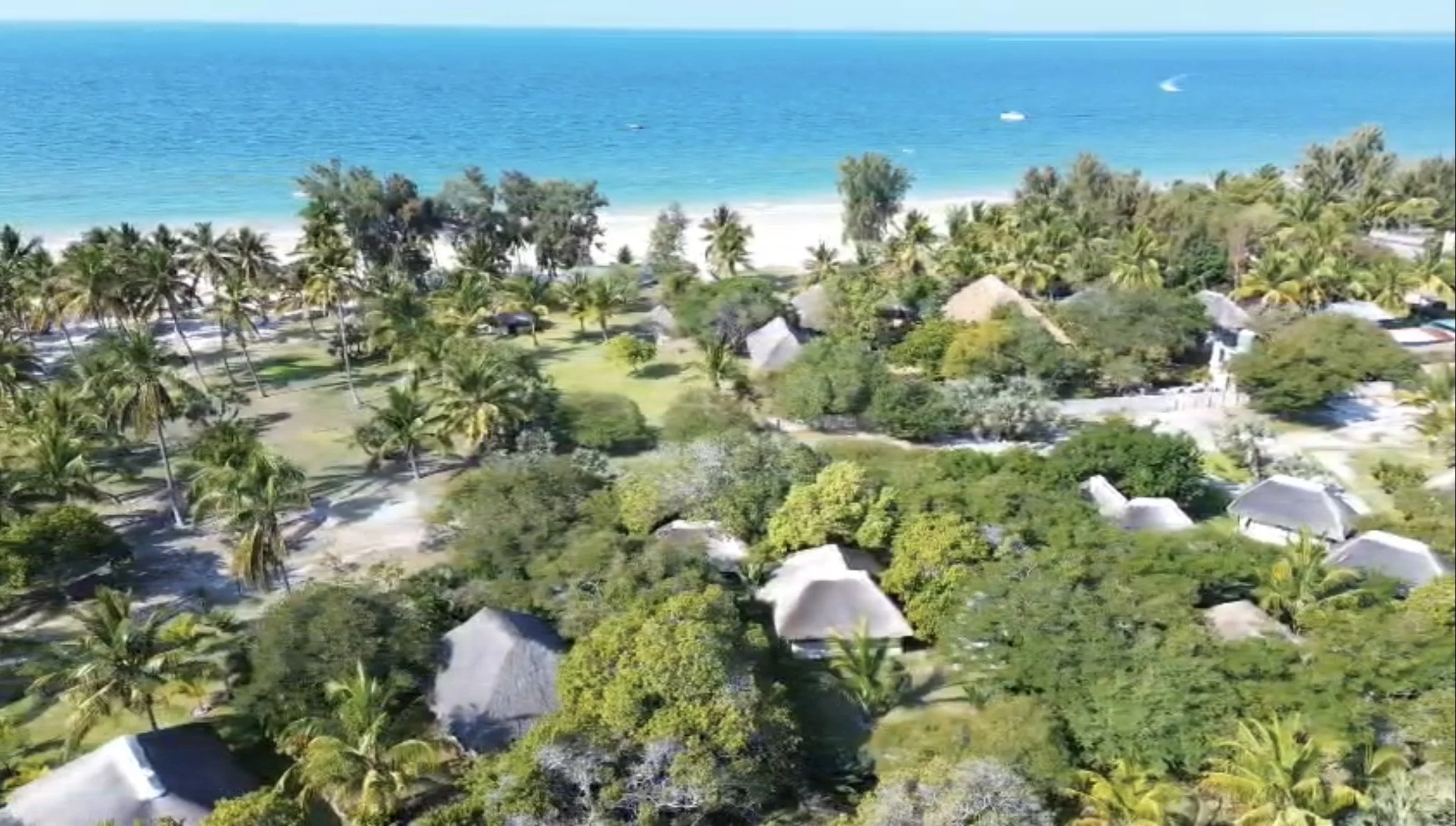 Aerial view of a tropical beach resort with overwater bungalows, lush green trees, and clear blue water in the background.