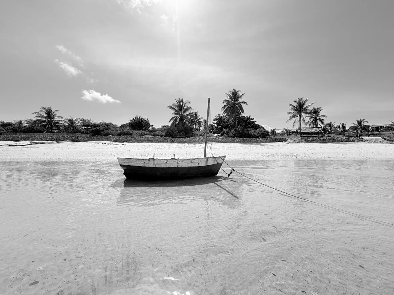 A small boat anchored on a calm sandy beach with palm trees and modest buildings in the background, under a partly cloudy sky.