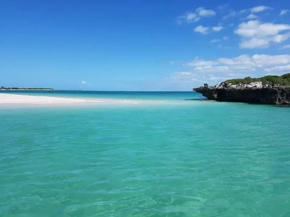Turquoise ocean waters with a sandy beach and rocky cliff in the background under a blue sky with scattered clouds.