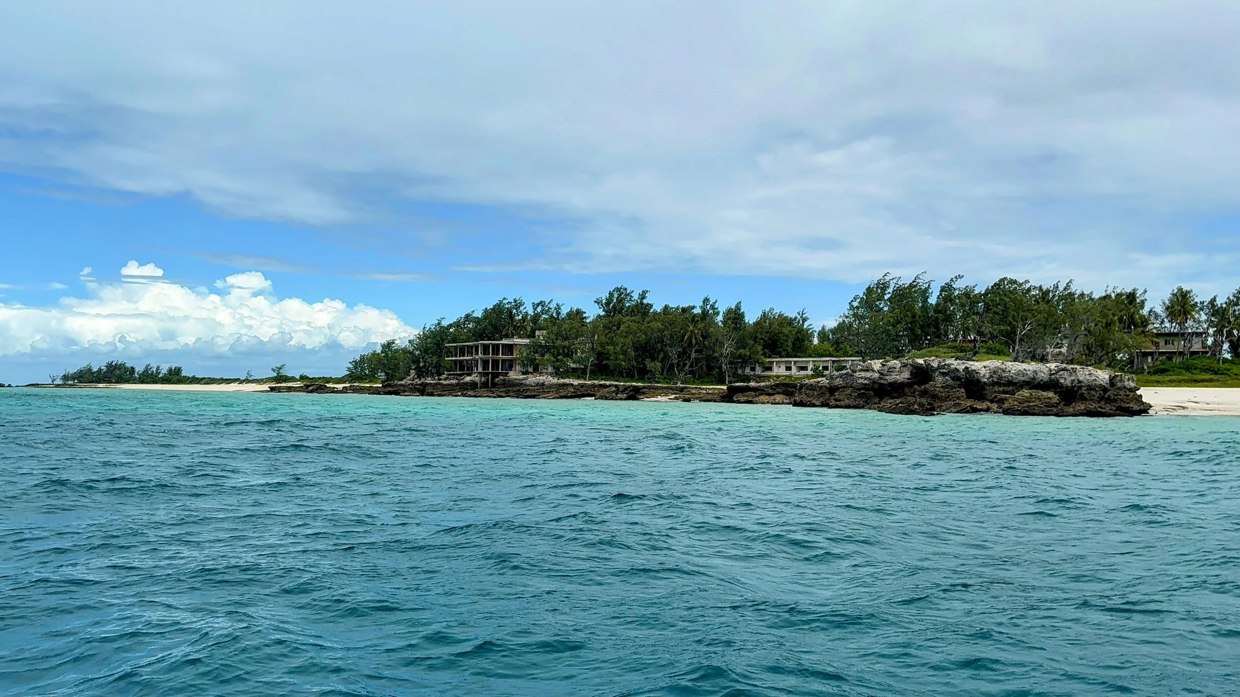 A view of a tropical shoreline with clear turquoise water in the foreground, rocky coastlines, lush green trees, and buildings on the hillside under a partly cloudy sky.