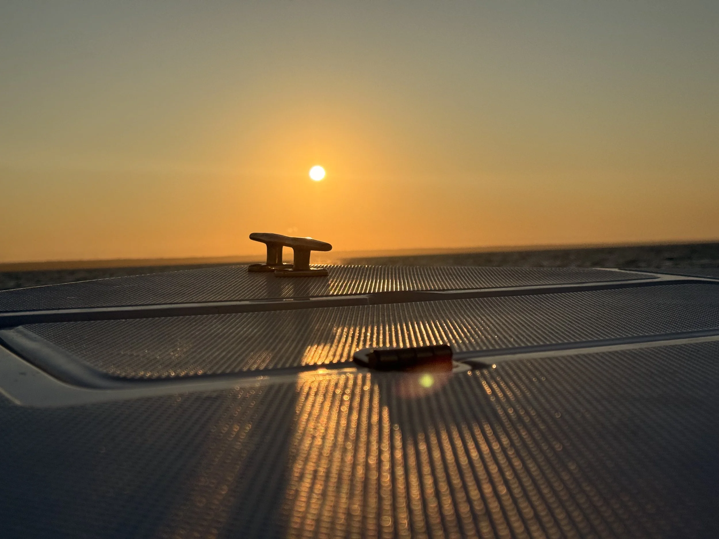 A sunset over the ocean as viewed from a boat or coast, with a metal cleat in the foreground on a dark surface, likely part of a boat deck.