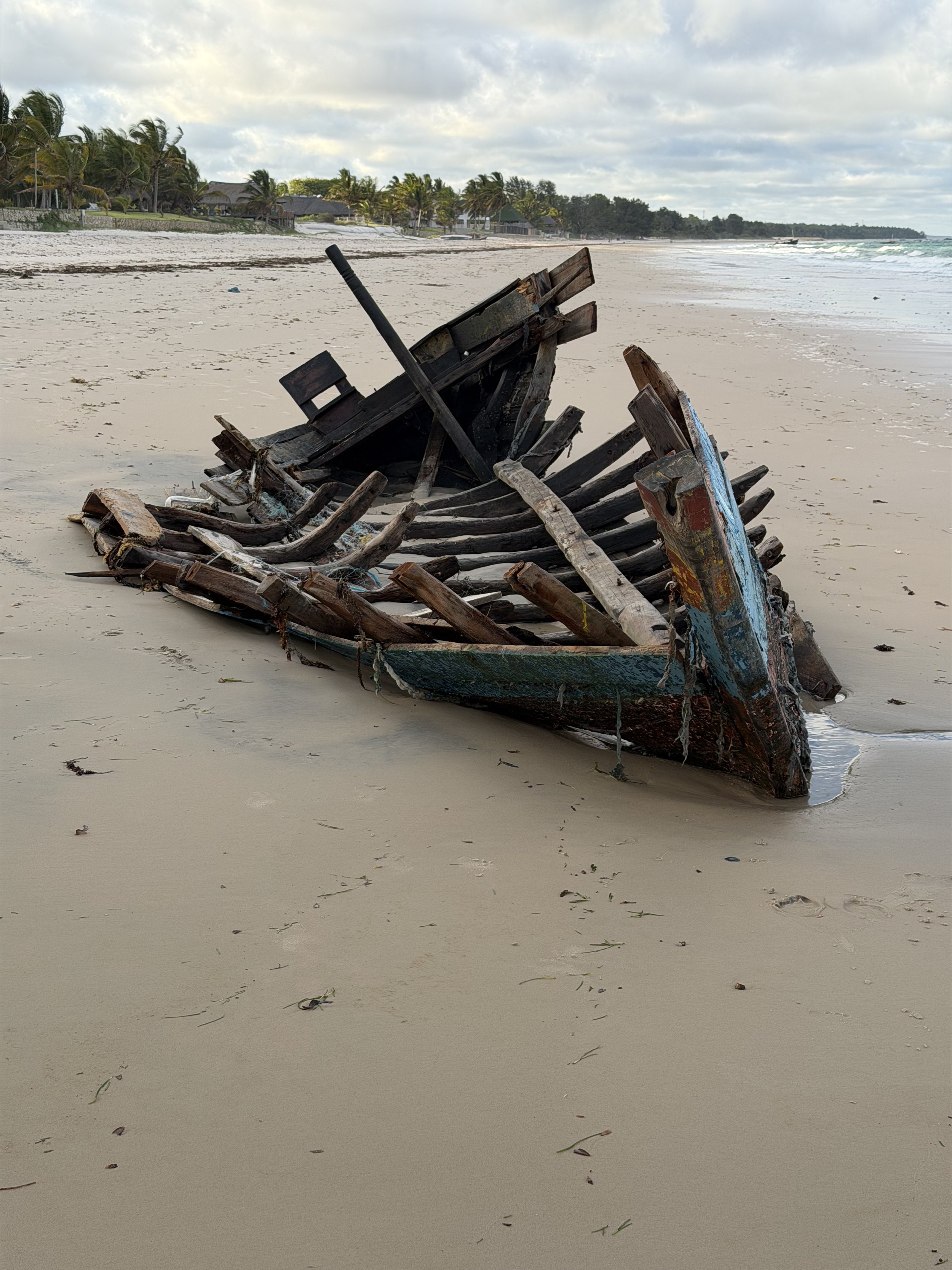 A weathered, broken wooden boat on a sandy beach with palm trees and buildings in the background under cloudy skies.
