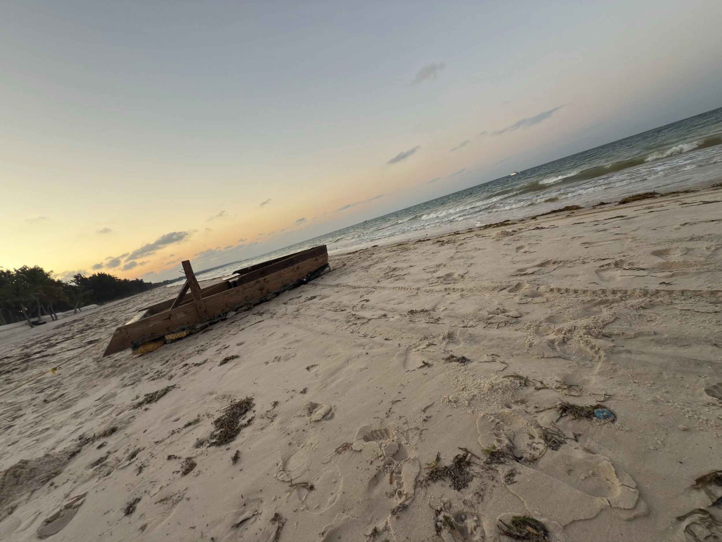 A sandy beach during sunset with a wooden boat on the shore, some trees in the background, and the ocean waves approaching the beach.