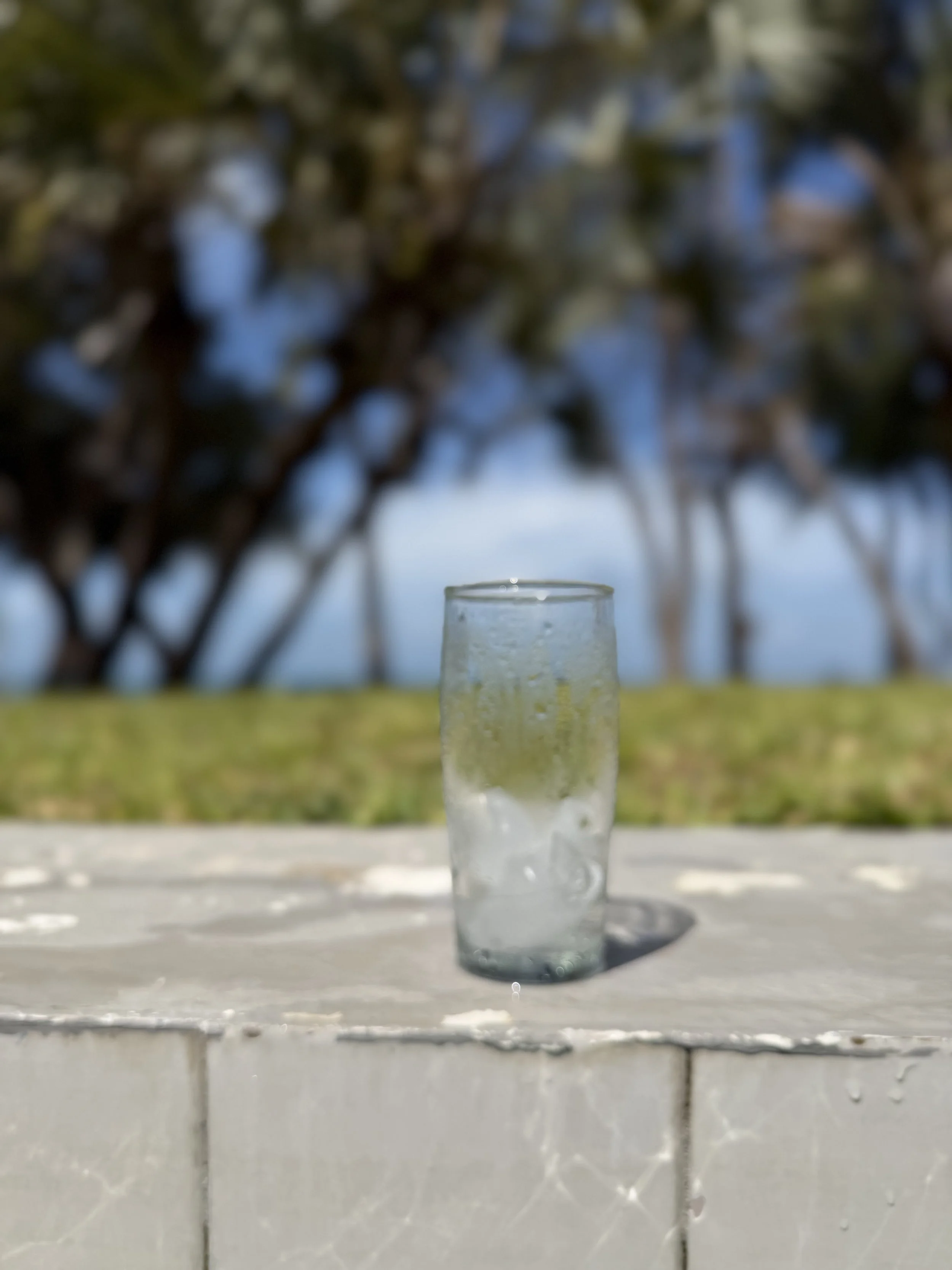 A clear glass filled with water placed on a white surface outdoors, with a blurred background of trees and sky.