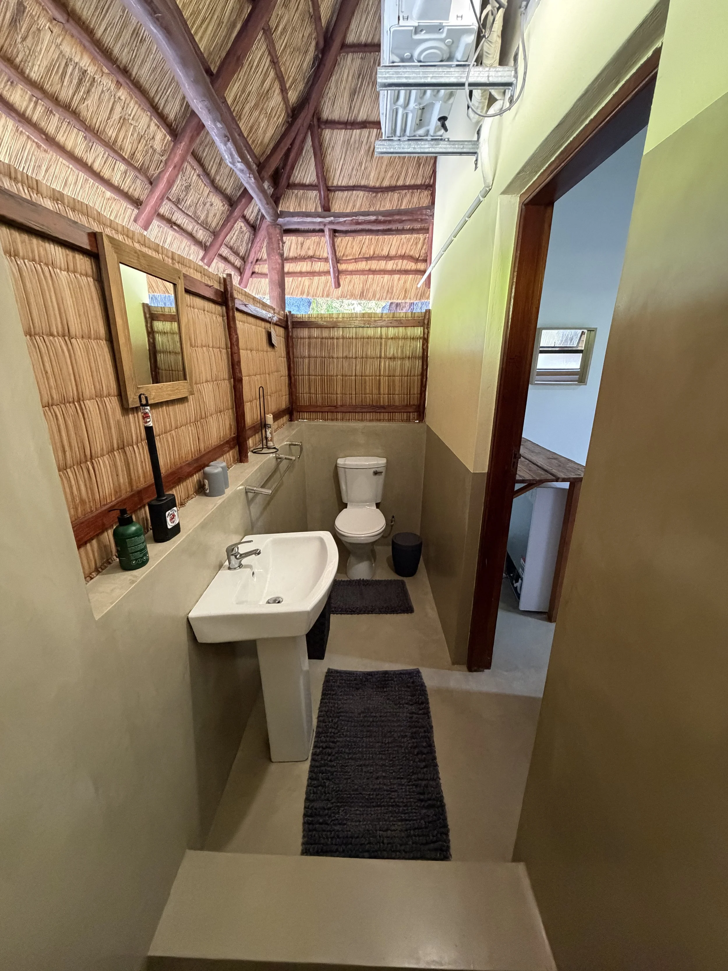 Bathroom with a small sink, toilet, black trash bin, mirror, and wooden wall panels made of bamboo, with a thatched roof ceiling and a window.