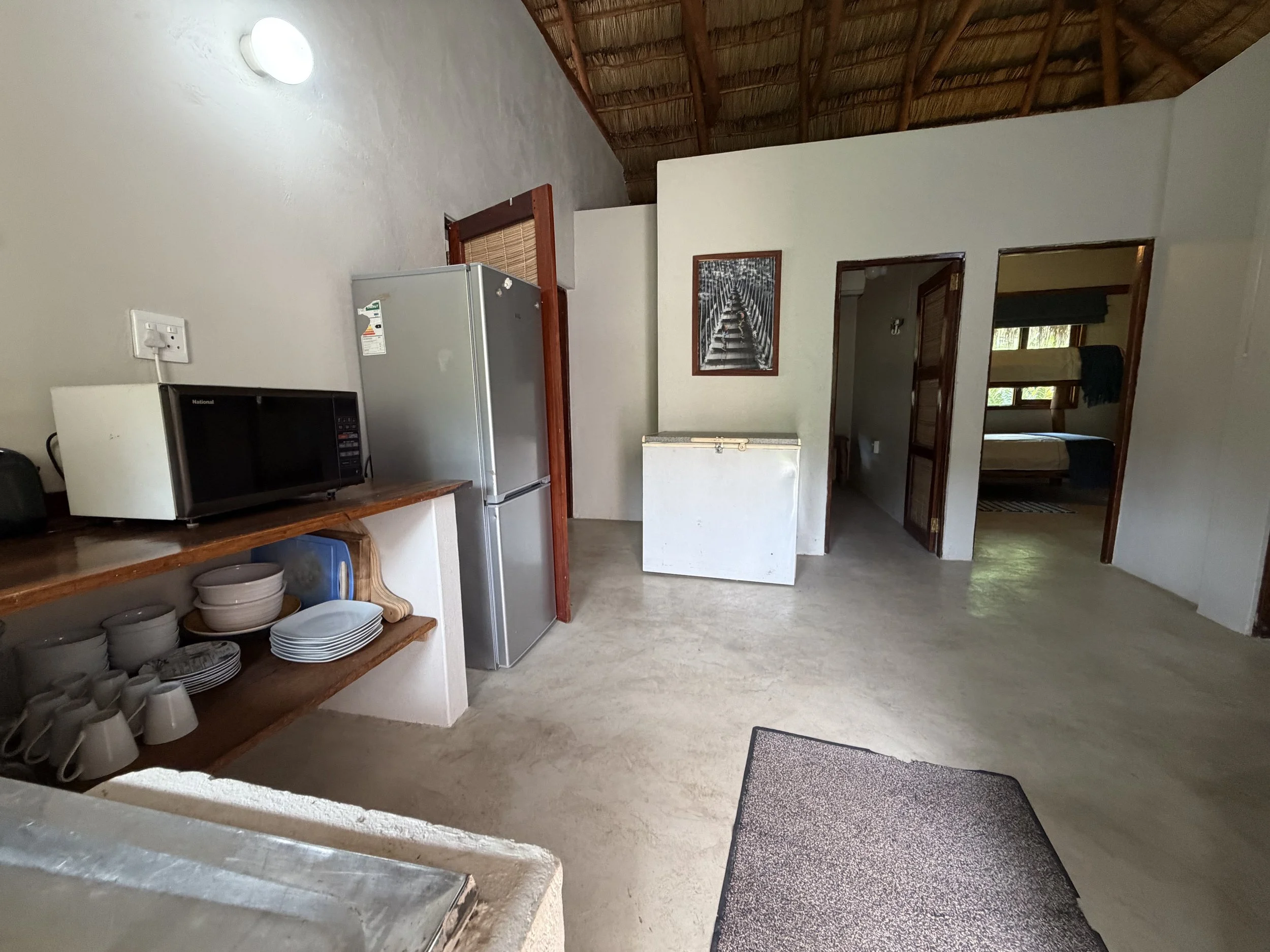 Interior of a room with a thatched roof, white walls, and an open doorway leading to a bedroom with a window and curtains. There is a small kitchen area with a microwave, refrigerator, a wooden shelf with dishes, and a wall picture of a bridge or walkway.