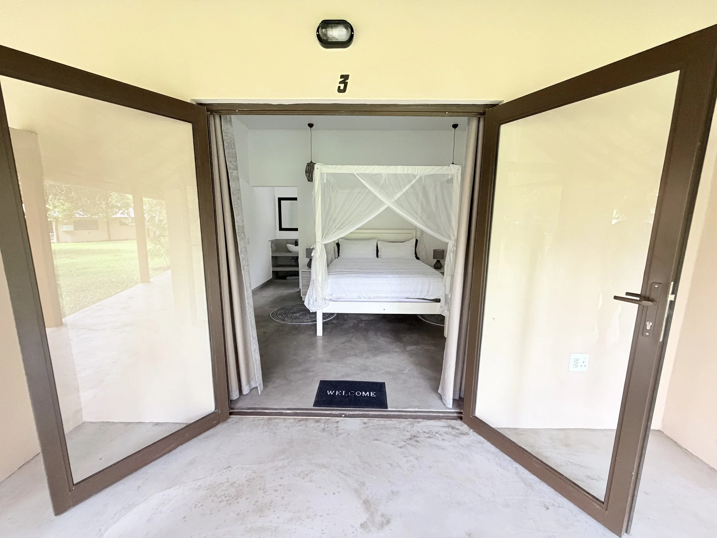 A bedroom with a white four-poster bed, a bedside table, and a wall mirror, viewed through open glass doors.