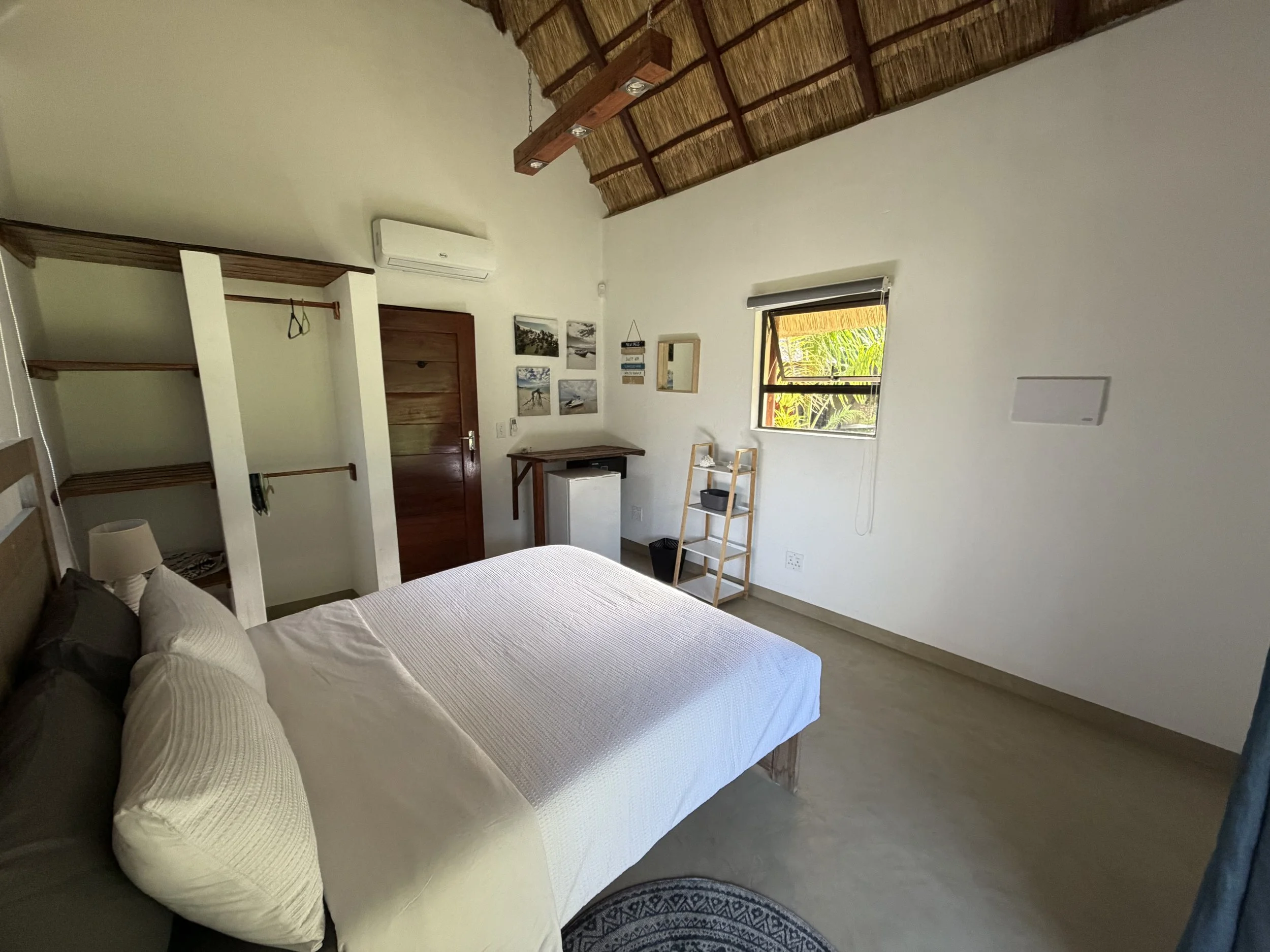 A bedroom with a large bed covered in white bedding, positioned next to a window with tropical greenery visible outside. The room features a high, thatched roof and walls decorated with framed pictures. A small desk and shelving unit are present, with a laundry basket underneath.