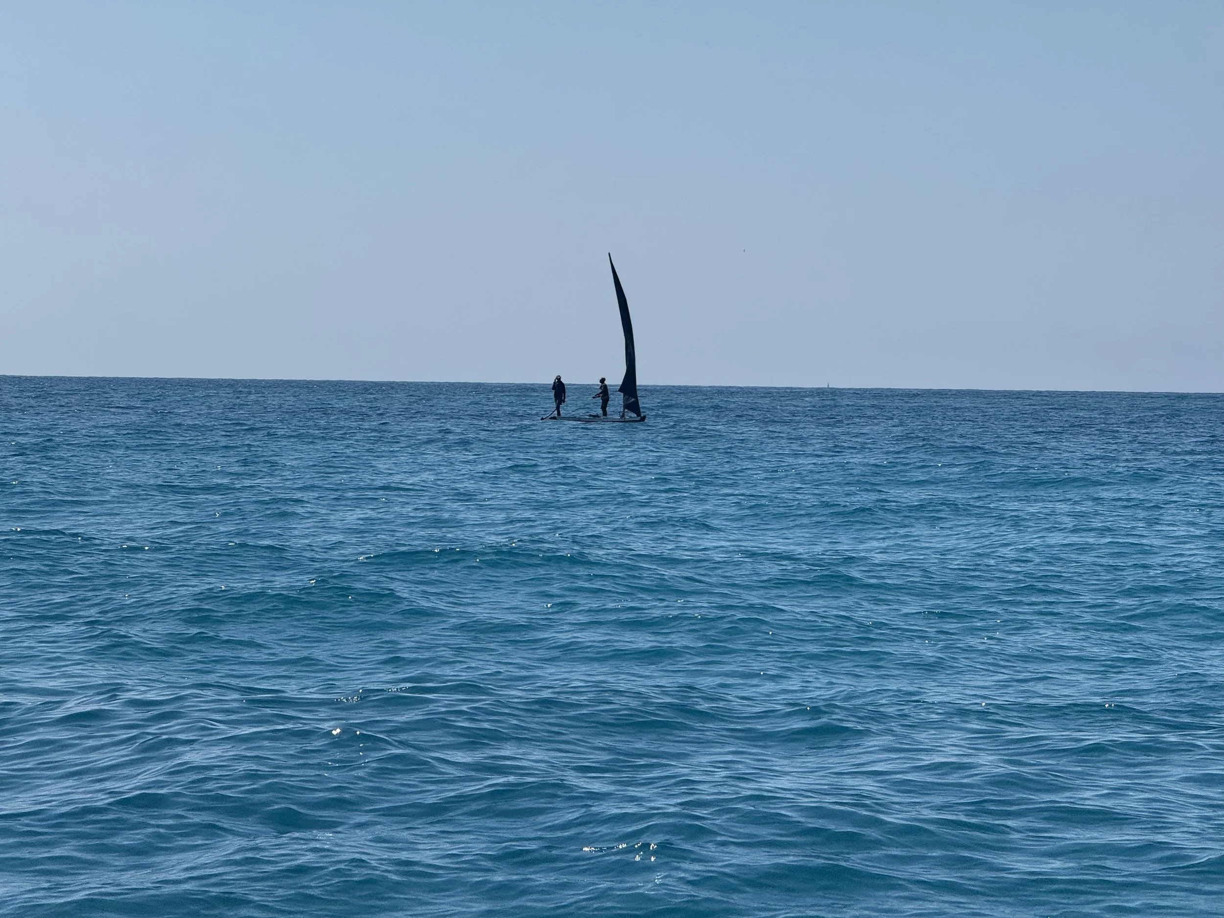 Two people stand on a small sailboat with a black sail, floating on the ocean with a clear blue sky.