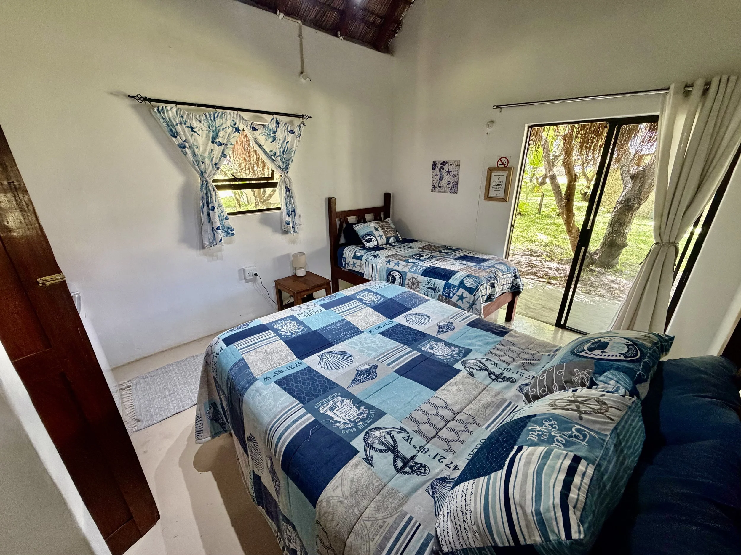 Two beds with blue and white nautical-themed bedspreads in a bright room with sliding glass door and window, showing outdoor trees and greenery.