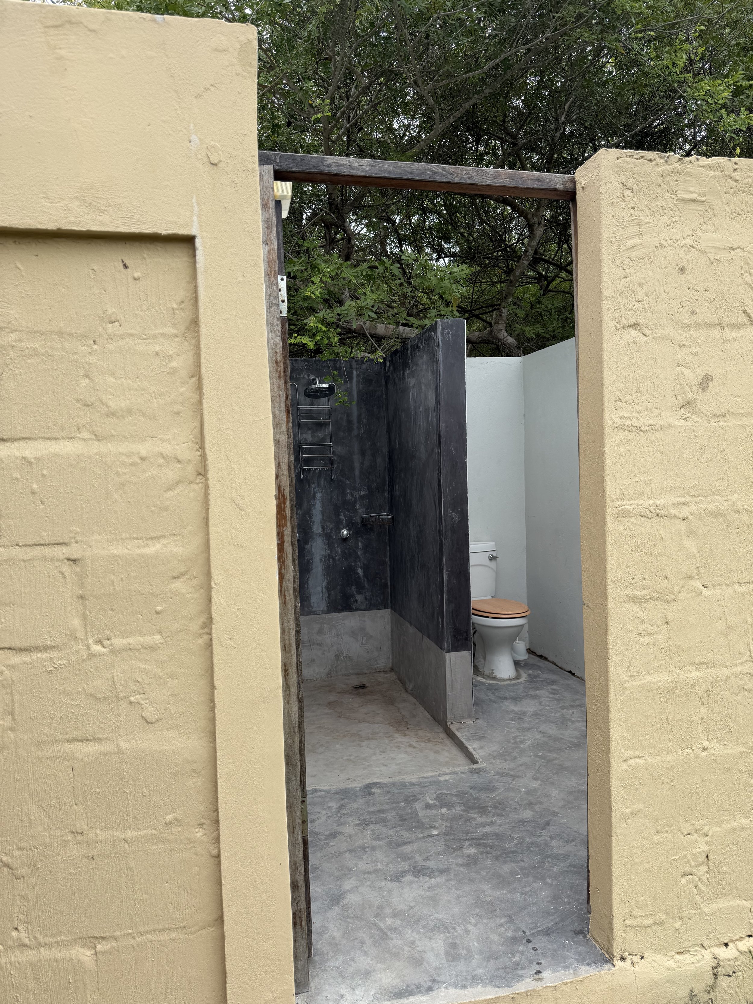 An outdoor bathroom with partial walls, a black toilet with a wooden seat, and a shower enclosure with a black wall and a hanging shower head, surrounded by trees and greenery in the background.