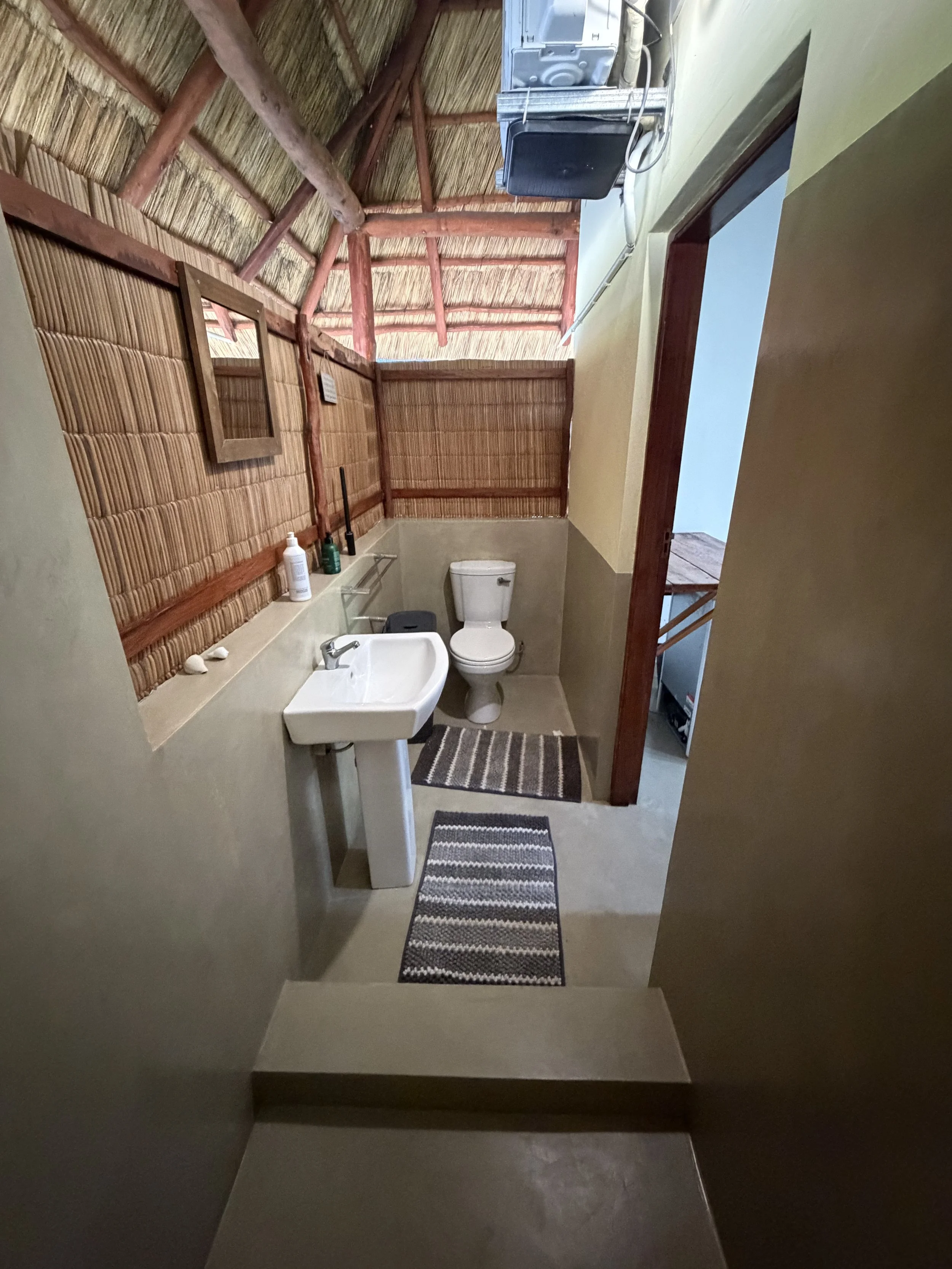 Bathroom with thatched roof ceiling, small sink, toilet, striped rugs, and a mirror on the wall.